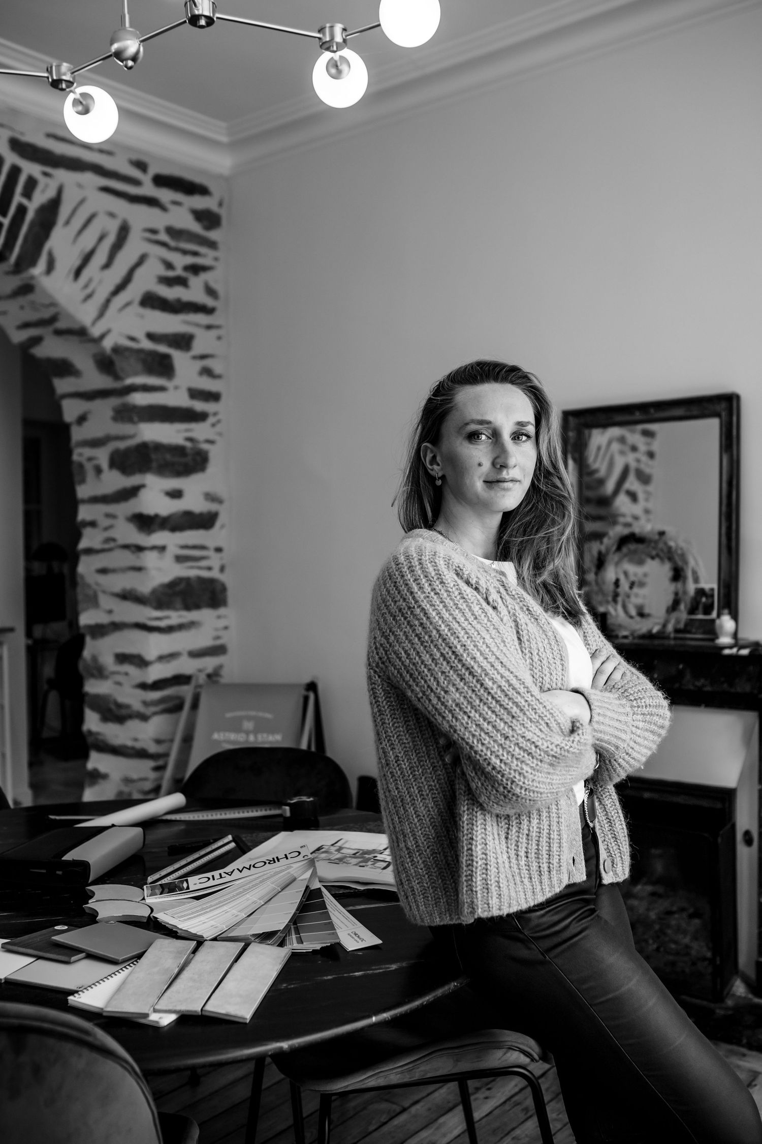 Black and white photo of someone in a knit sweater leaning against a desk in a room with stone walls.