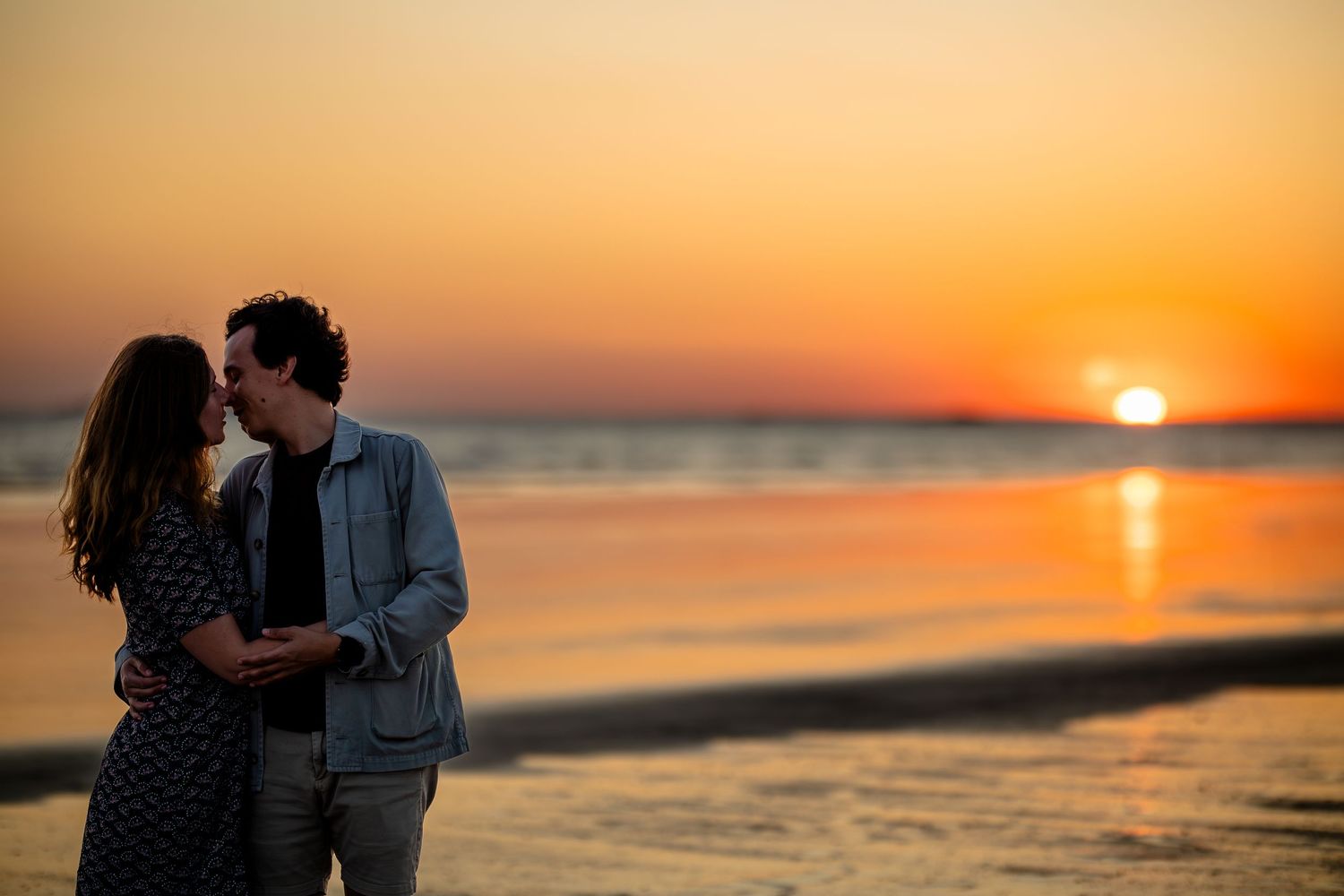 A couple embraces on a beach at sunset with the orange sun reflecting on calm ocean waters.