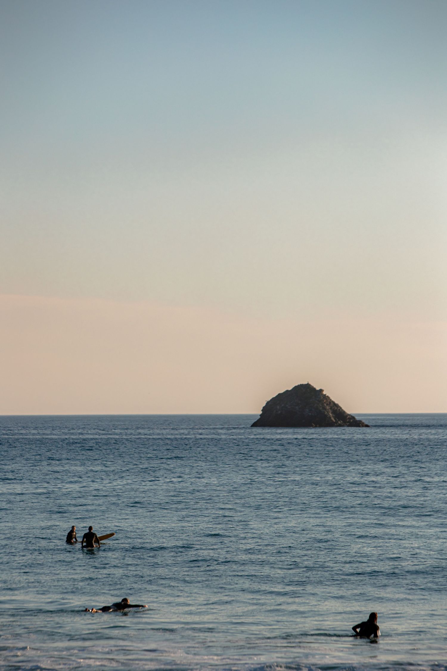 Surfers floating in calm ocean waters near a rocky island during sunset.
