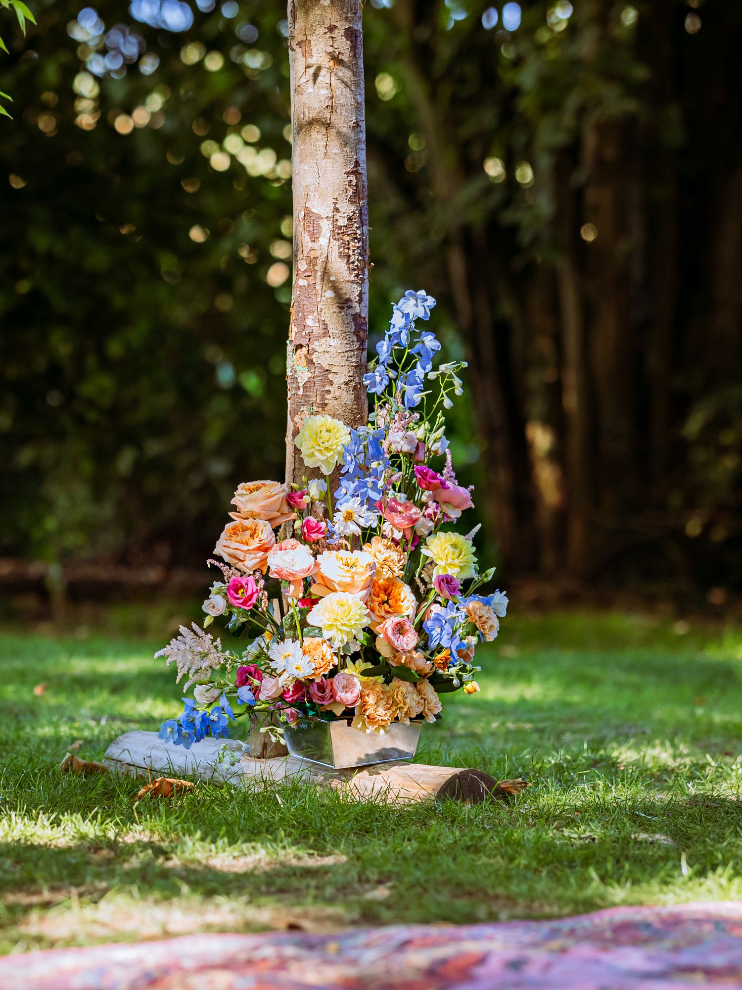 A colorful floral arrangement with blue and orange blooms sits at the base of a birch tree on green grass.