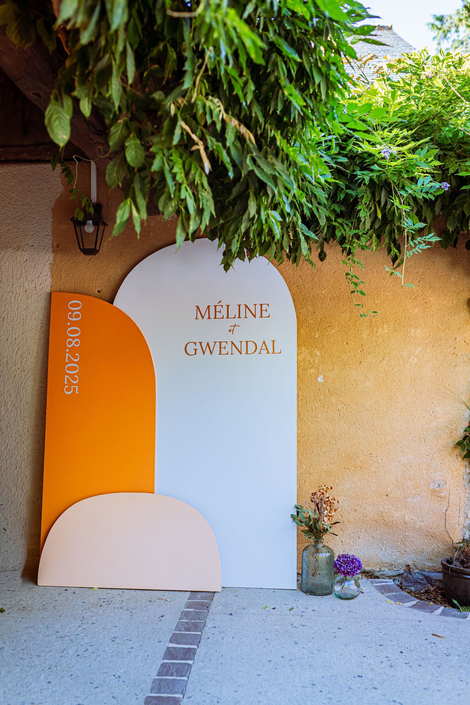Modern white and orange arched wedding signage with names displayed under lush green foliage against a beige wall.
