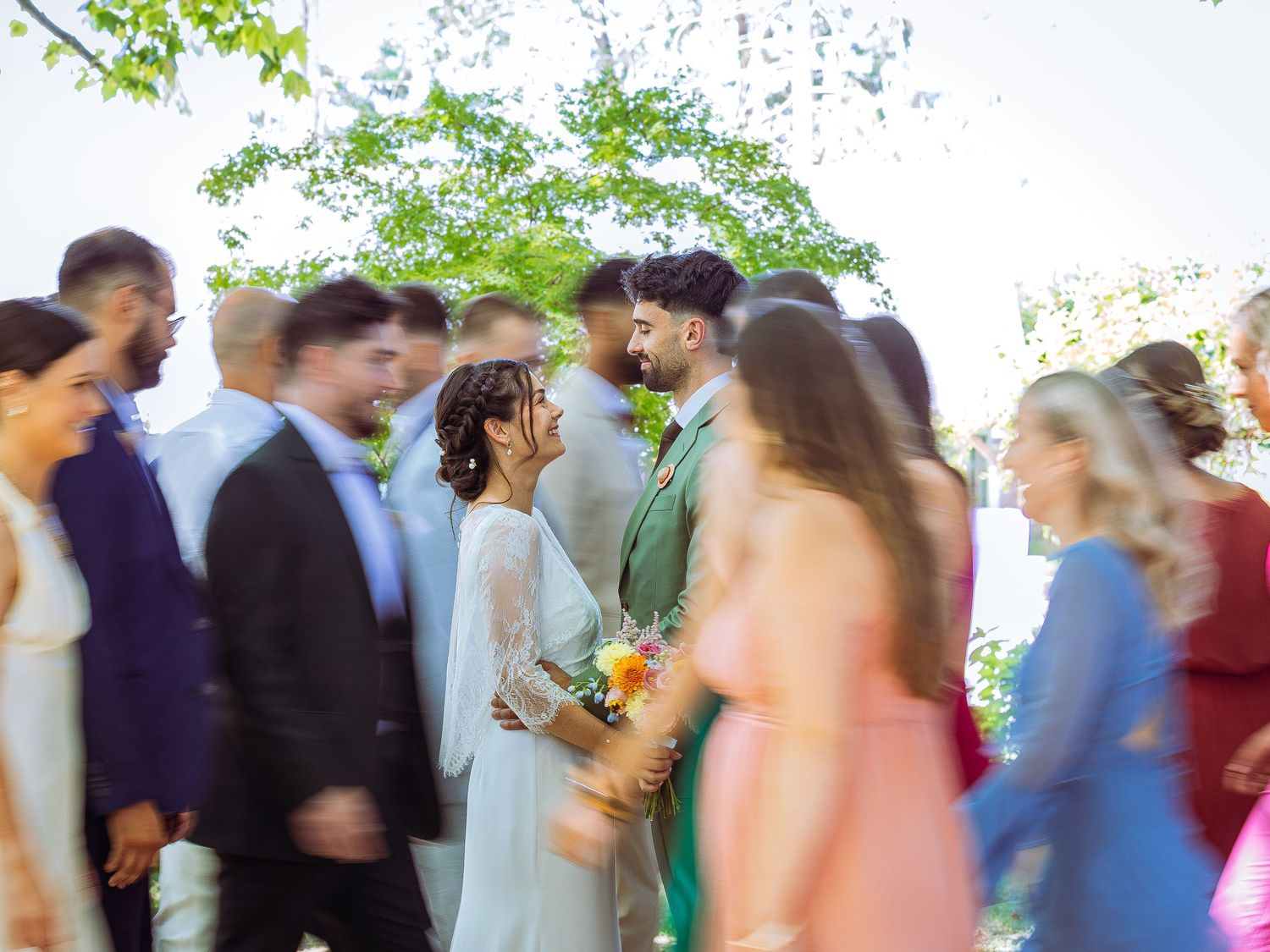 Blurred motion of wedding guests dancing and celebrating at an outdoor reception under green trees.
