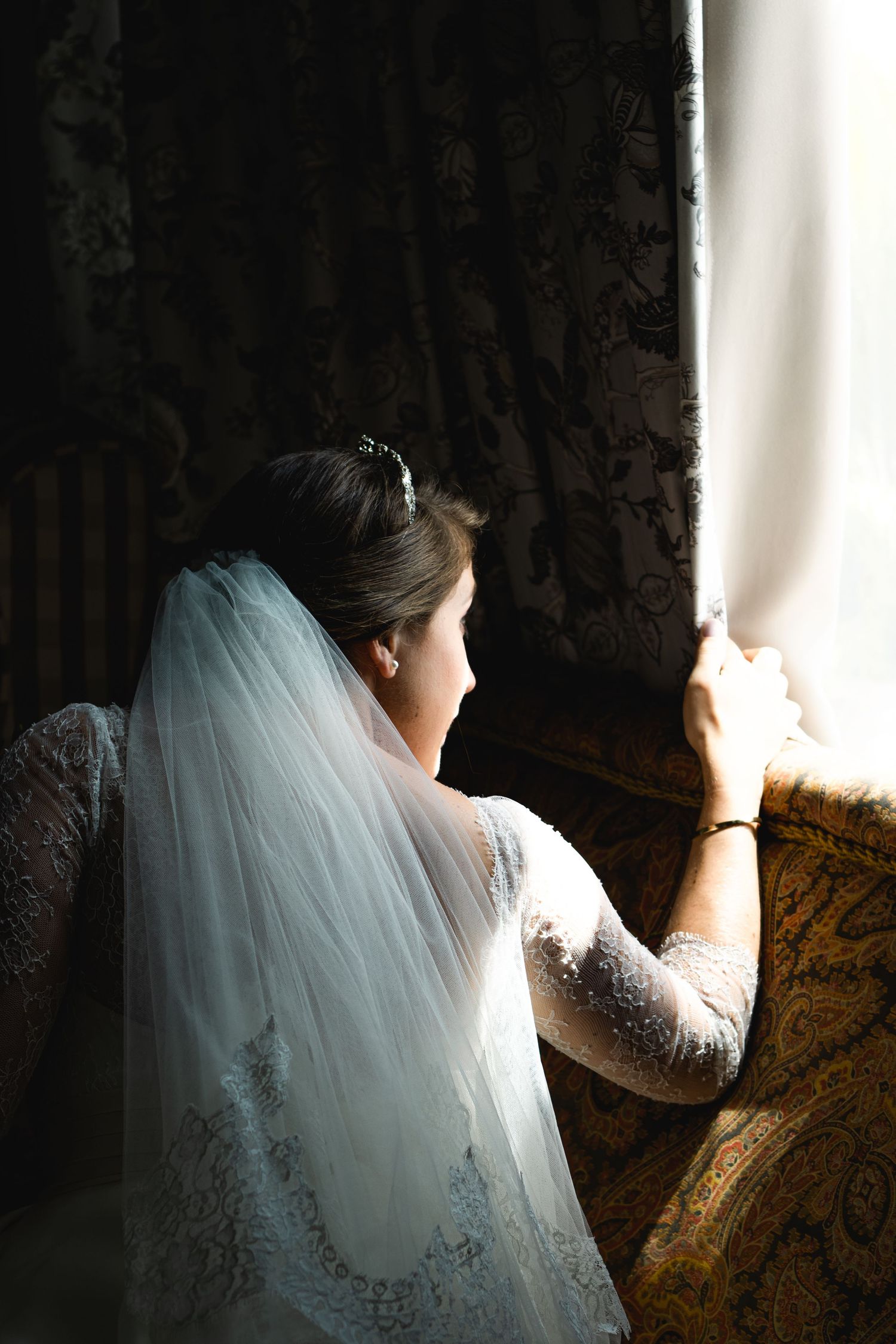 A bride in a white veil gazes through a window in dramatic side lighting.