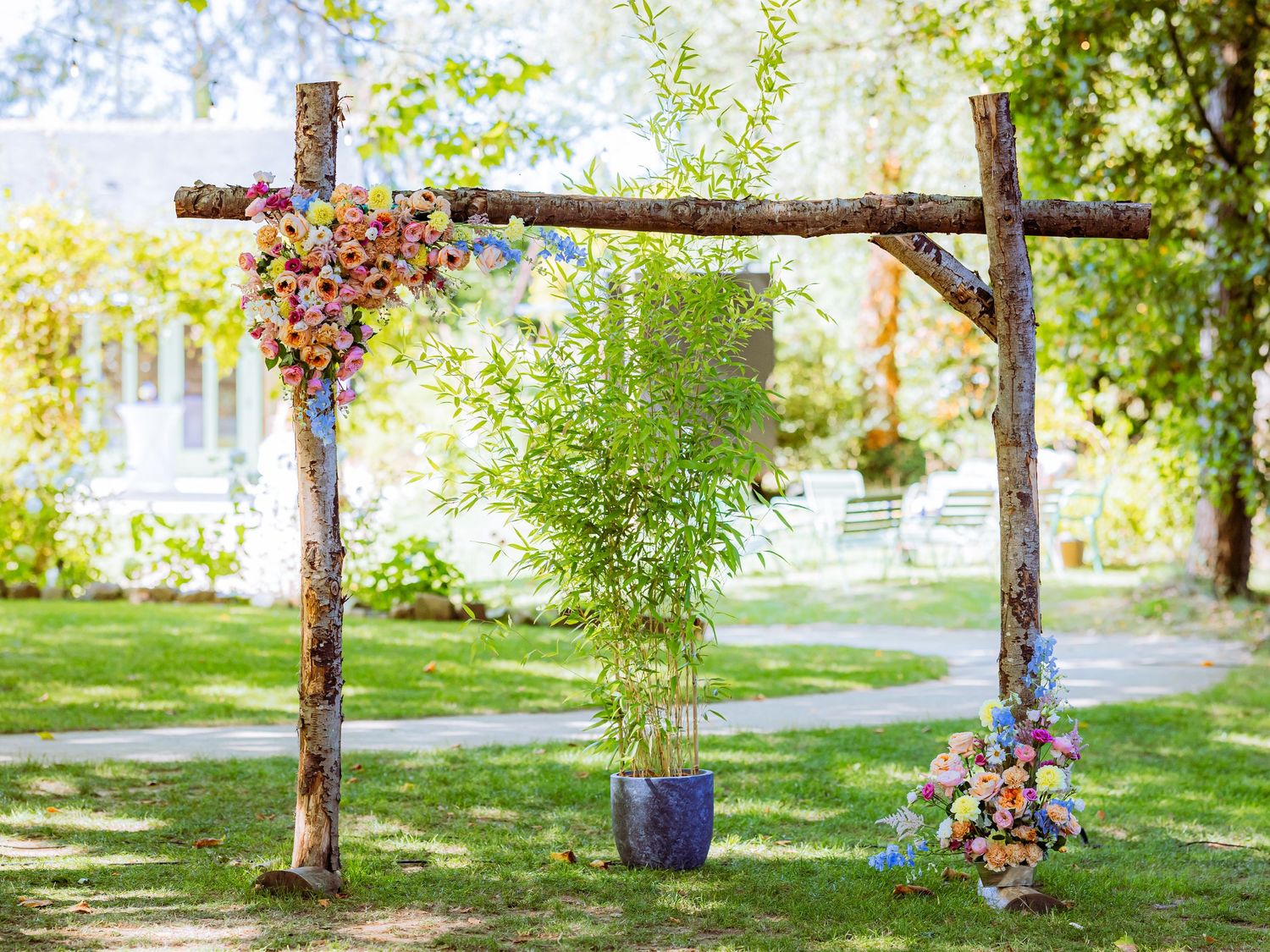A rustic wooden wedding arch decorated with colorful flowers stands in a sunny garden setting.