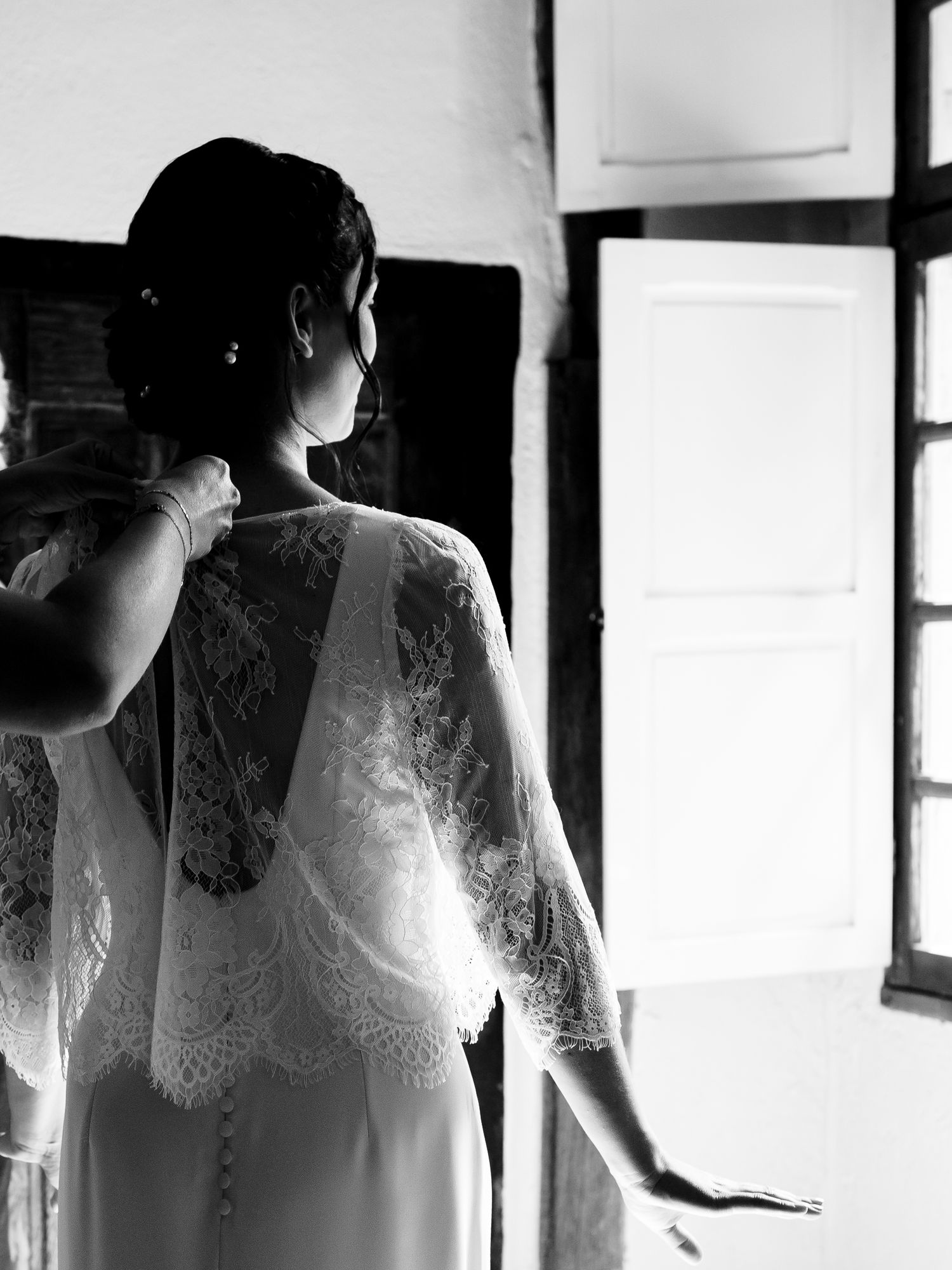 Black and white photo of someone wearing a delicate lace wedding dress with buttons and long sleeves by a window.