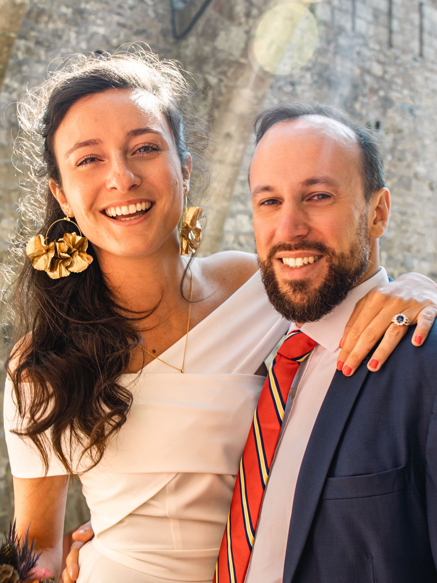 Newlywed couple in formal attire smiling against stone wall background.