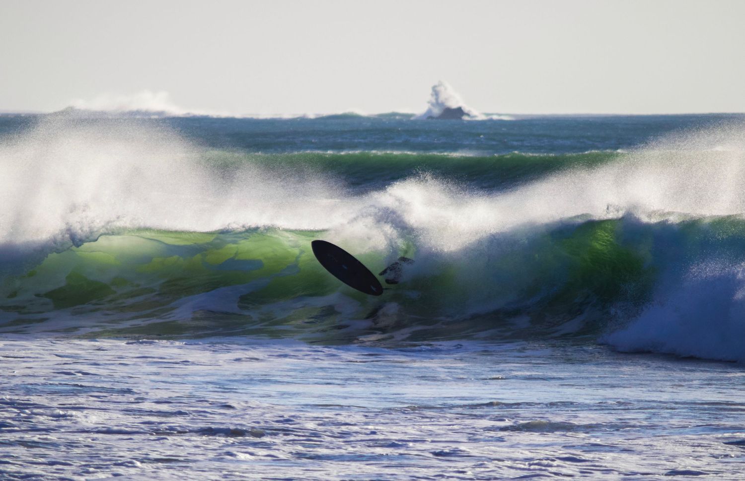 A surfer rides through a massive emerald wave with a lighthouse visible in the background.