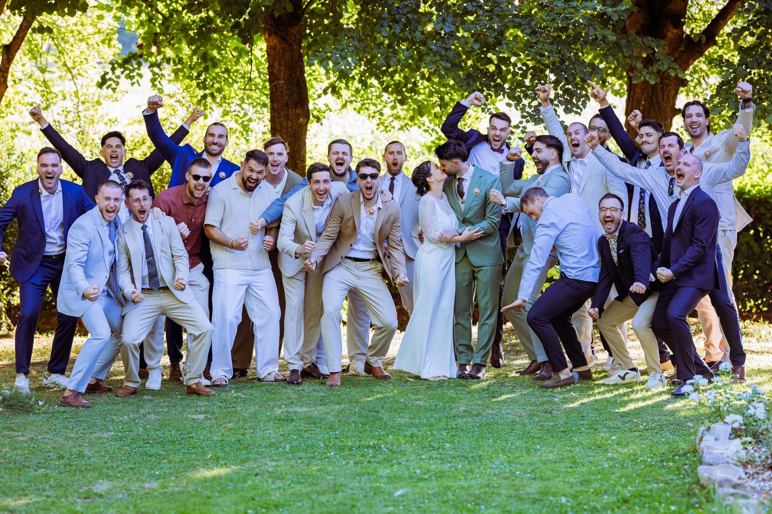 Wedding party in white and light colored attire poses playfully on grass under trees during outdoor celebration.