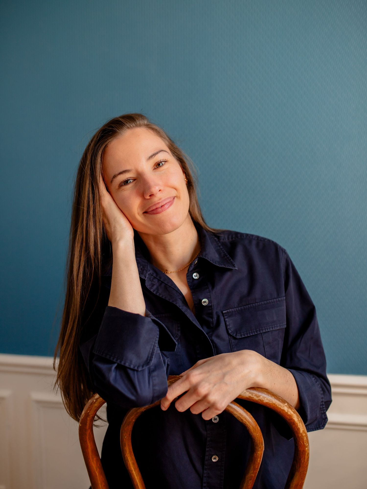 Casual portrait leaning on wooden chair with warm expression against blue wall.