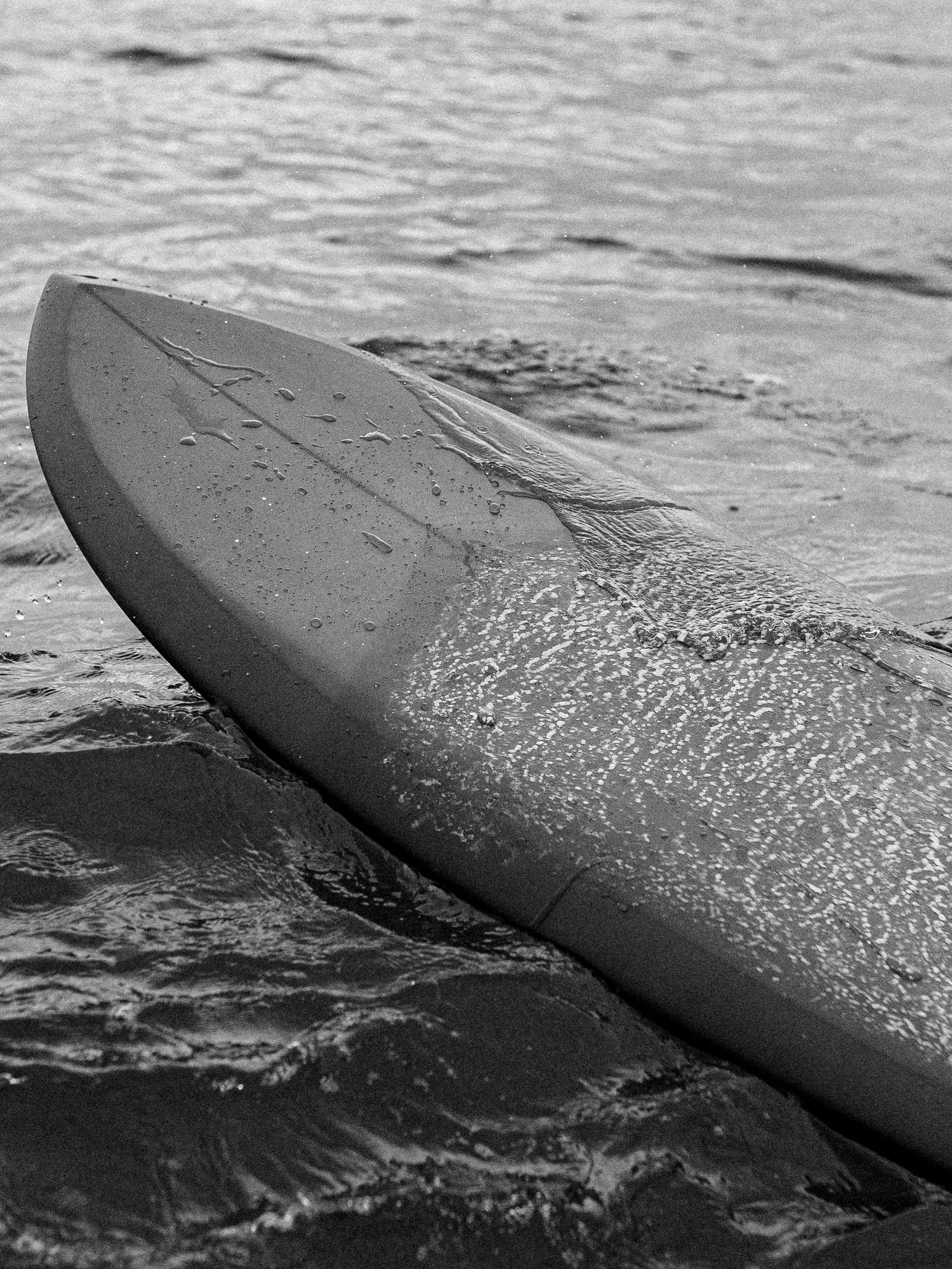 Black and white close-up photograph of a surfboard gliding through rippling ocean water.