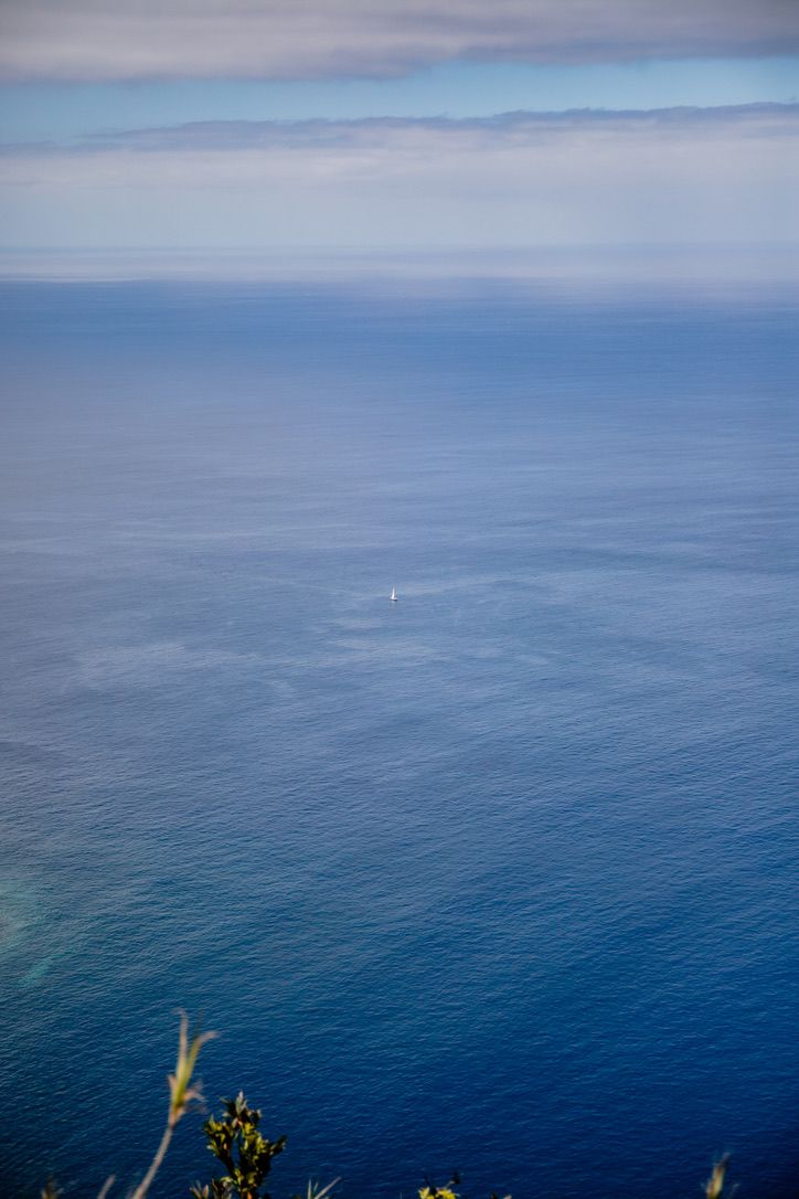 A lone white sailboat dots the vast blue ocean horizon against misty mountains in the distance.
