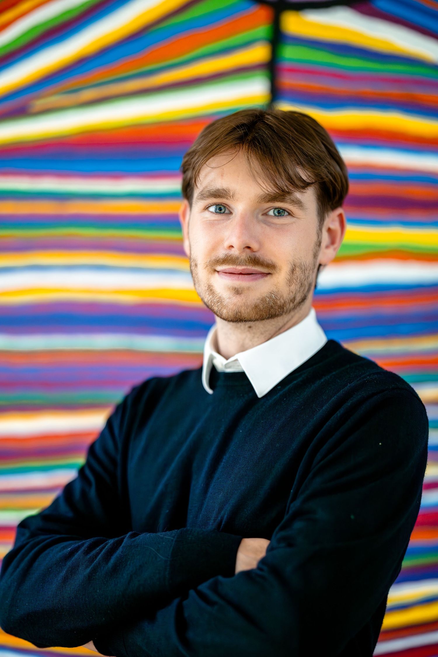 Professional portrait against a colorful striped background wearing a navy sweater and white collar.