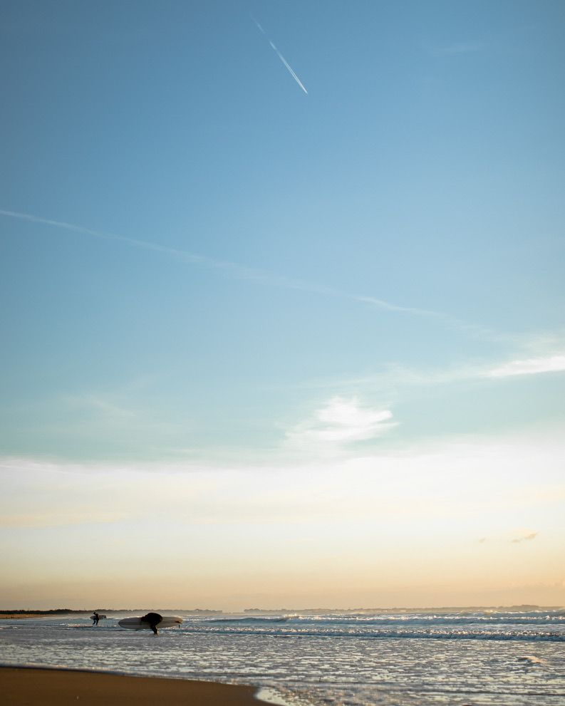 Peaceful beach sunset with contrails streaking across a pale blue sky over the ocean.