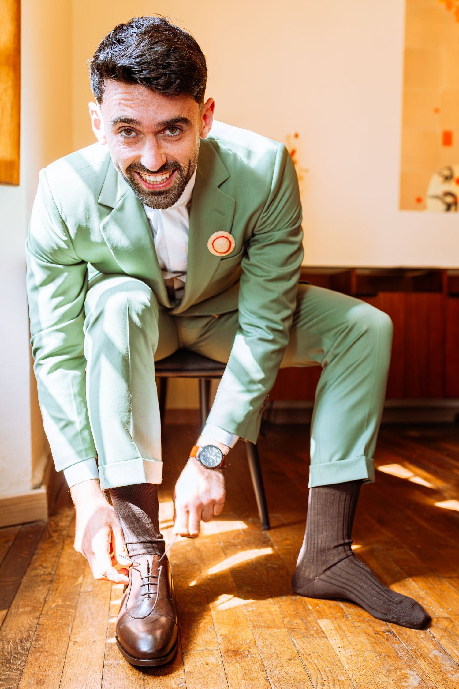 Someone in a mint green suit smiles while putting on brown leather dress shoes on a hardwood floor.