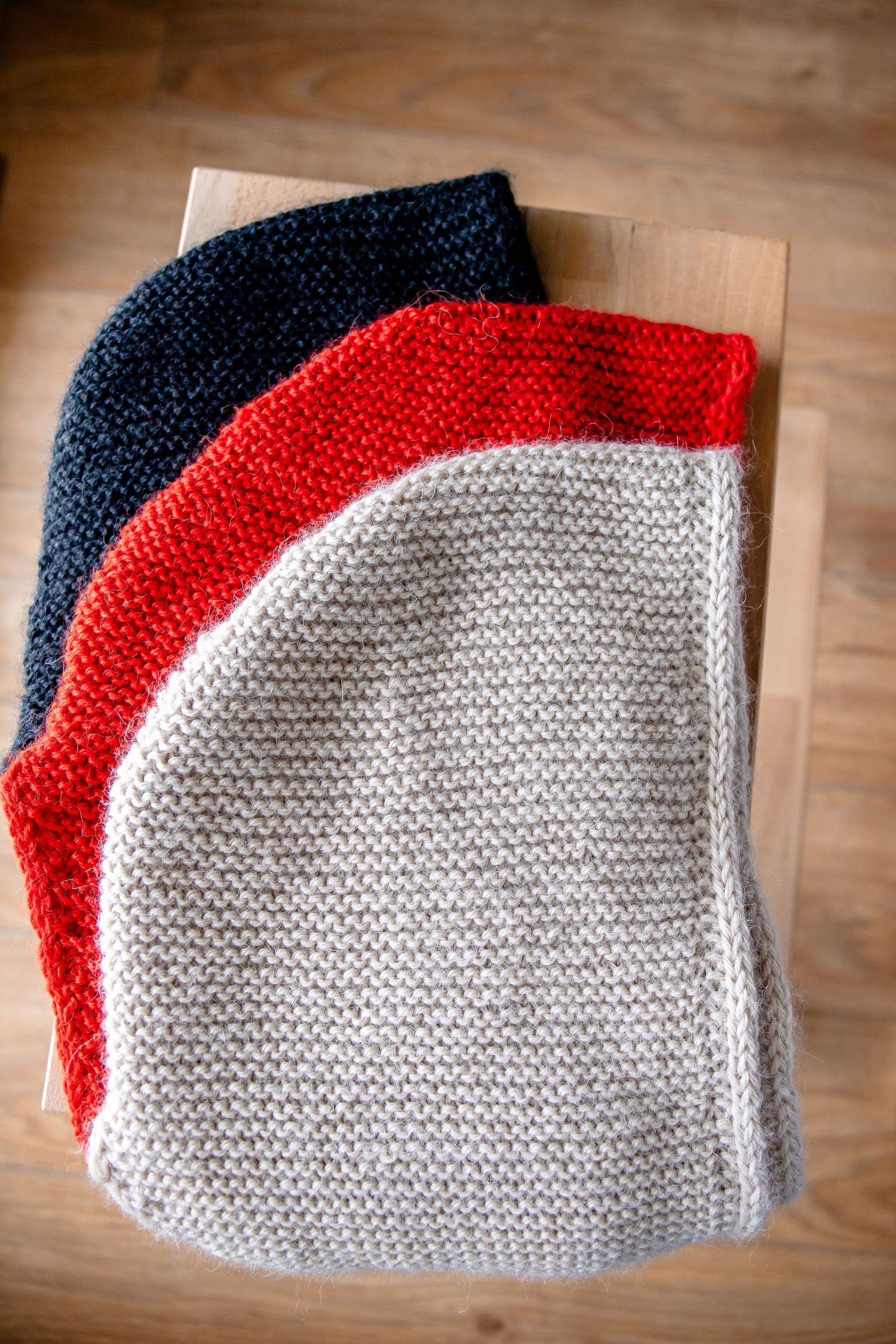 Knitted textiles in white, red, and navy arranged in a flat lay pattern on wooden flooring.
