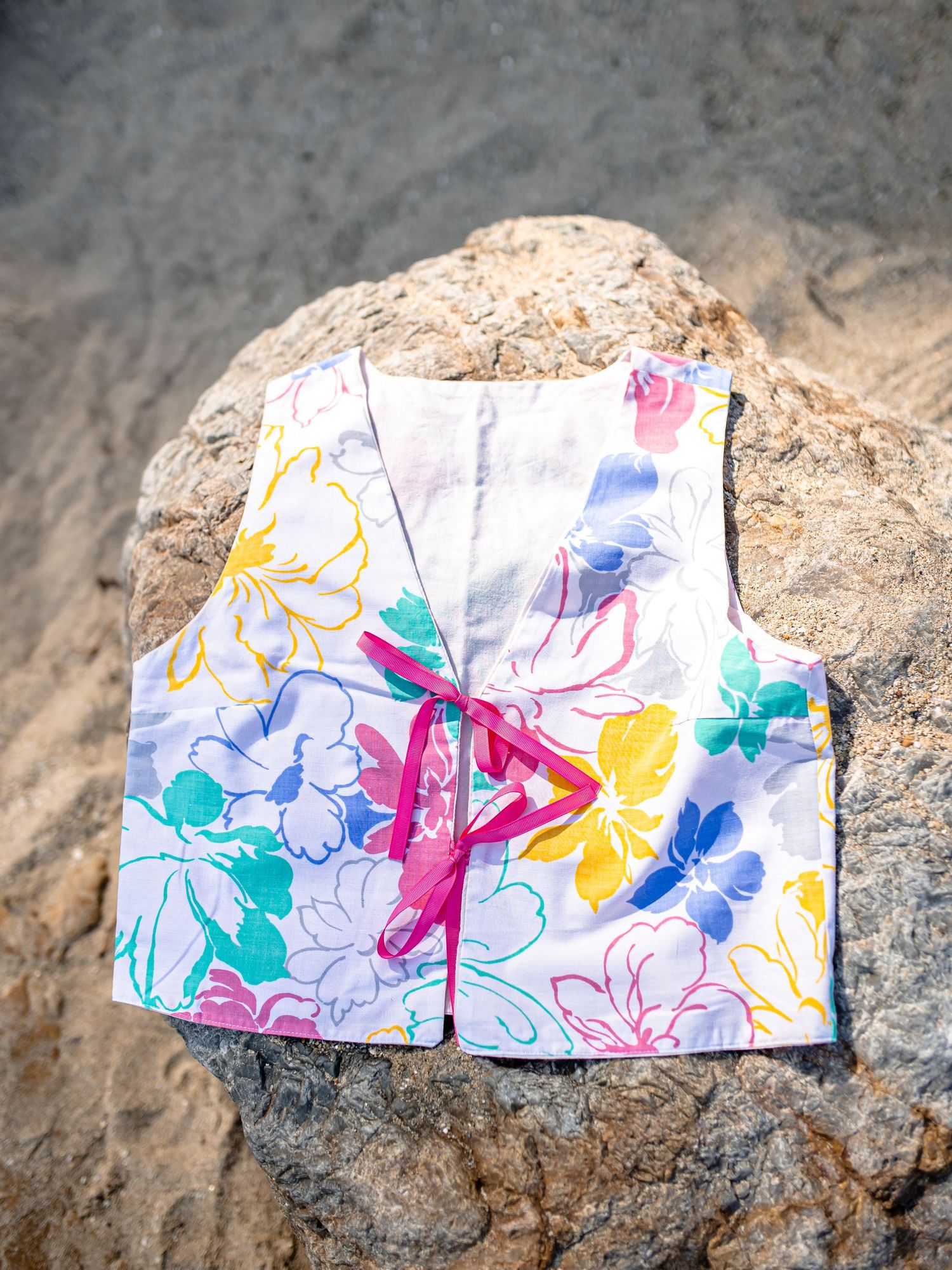 A colorful floral print baby vest with pink ribbon tie lies on a rocky surface.