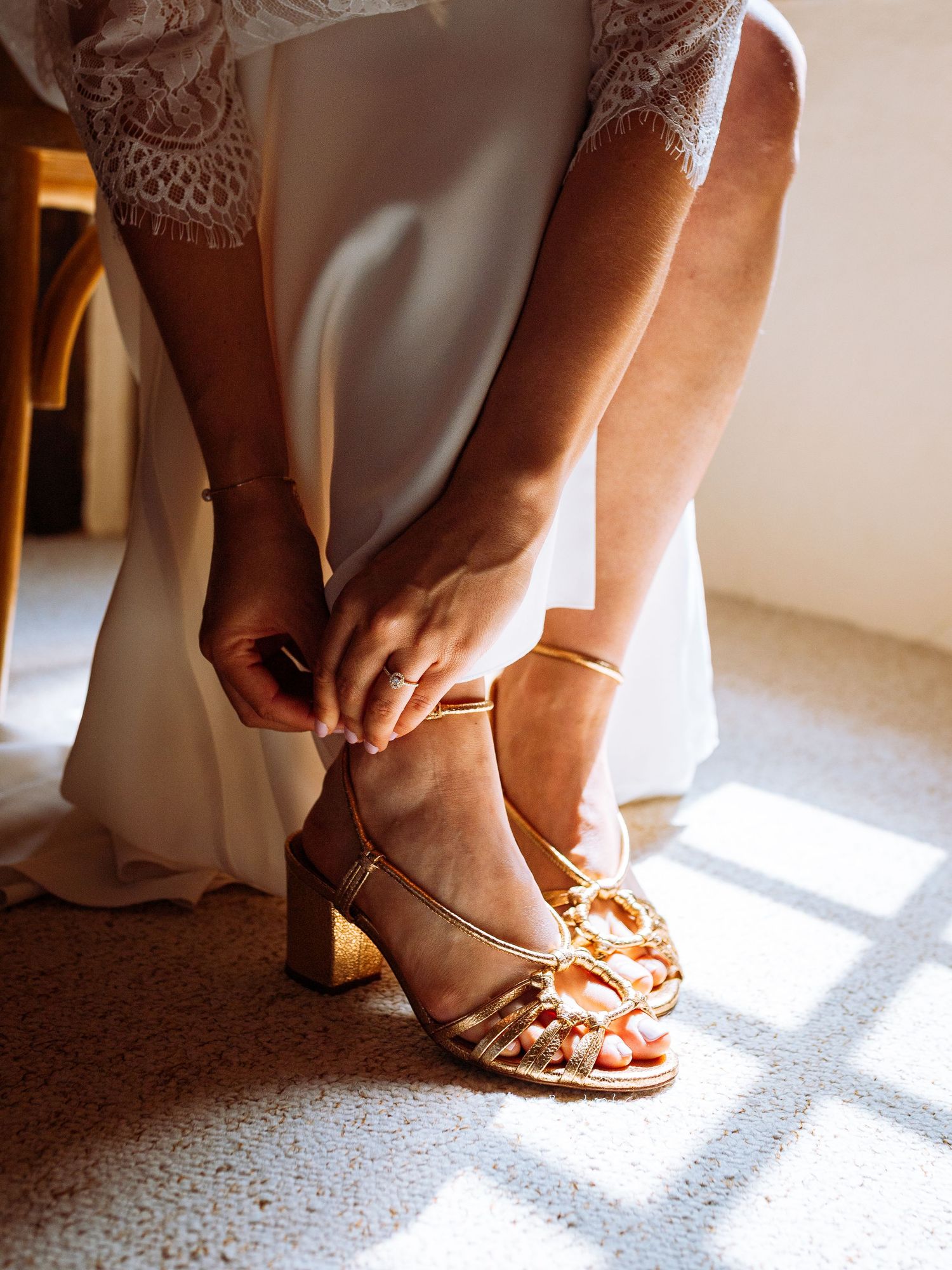 Close-up view of a bride putting on elegant gold wedding shoes in natural window light.