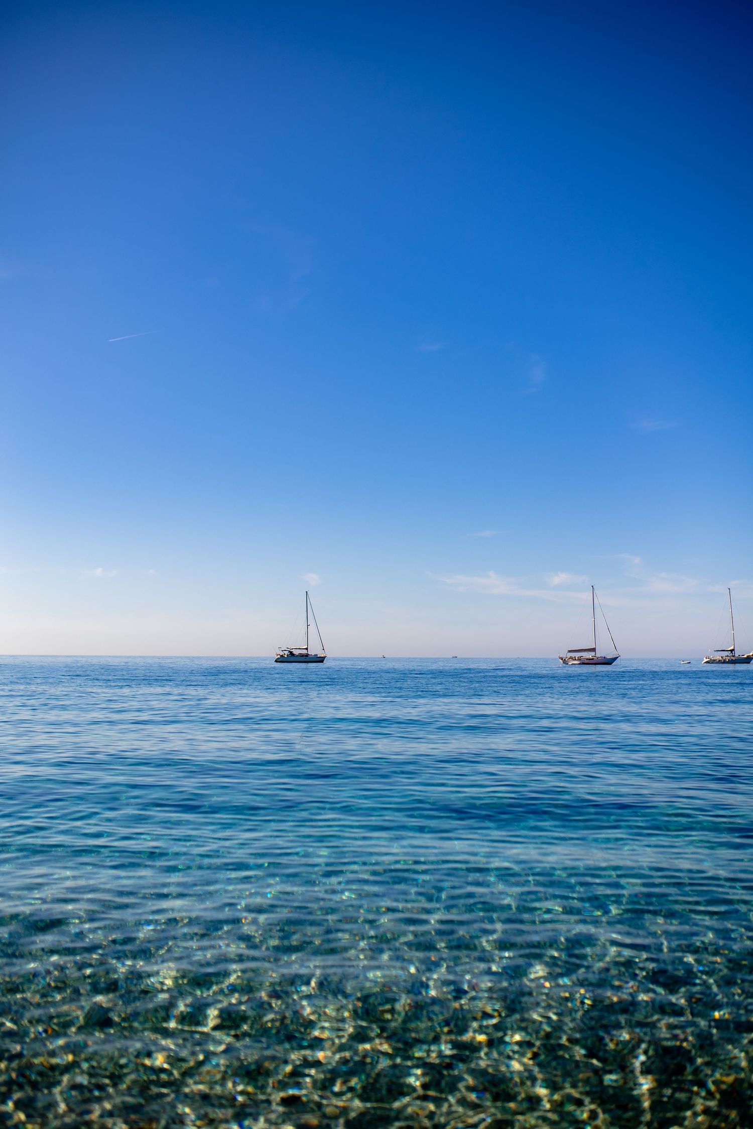 Crystal clear Mediterranean waters with sailboats anchored on the horizon against a vibrant blue sky.