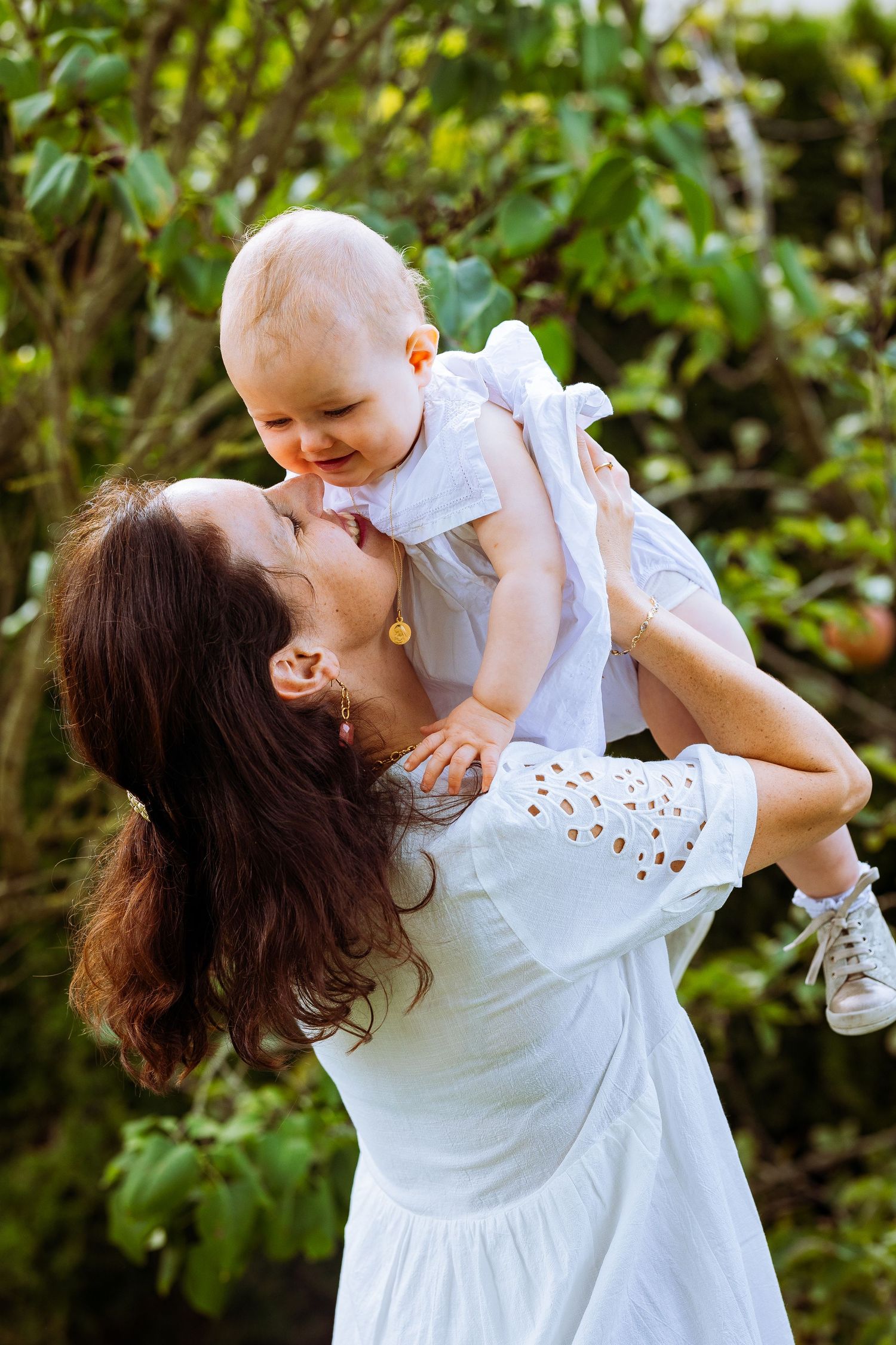 A tender moment between parent and baby in white outfits playing outdoors against lush green foliage.