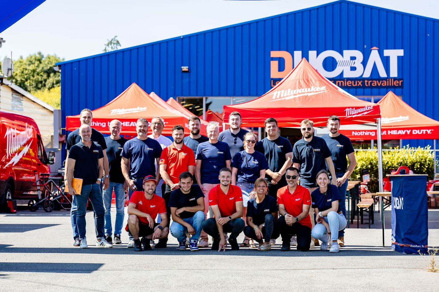 Group gathered in front of a blue building with Duobat branding and red canopy tents.