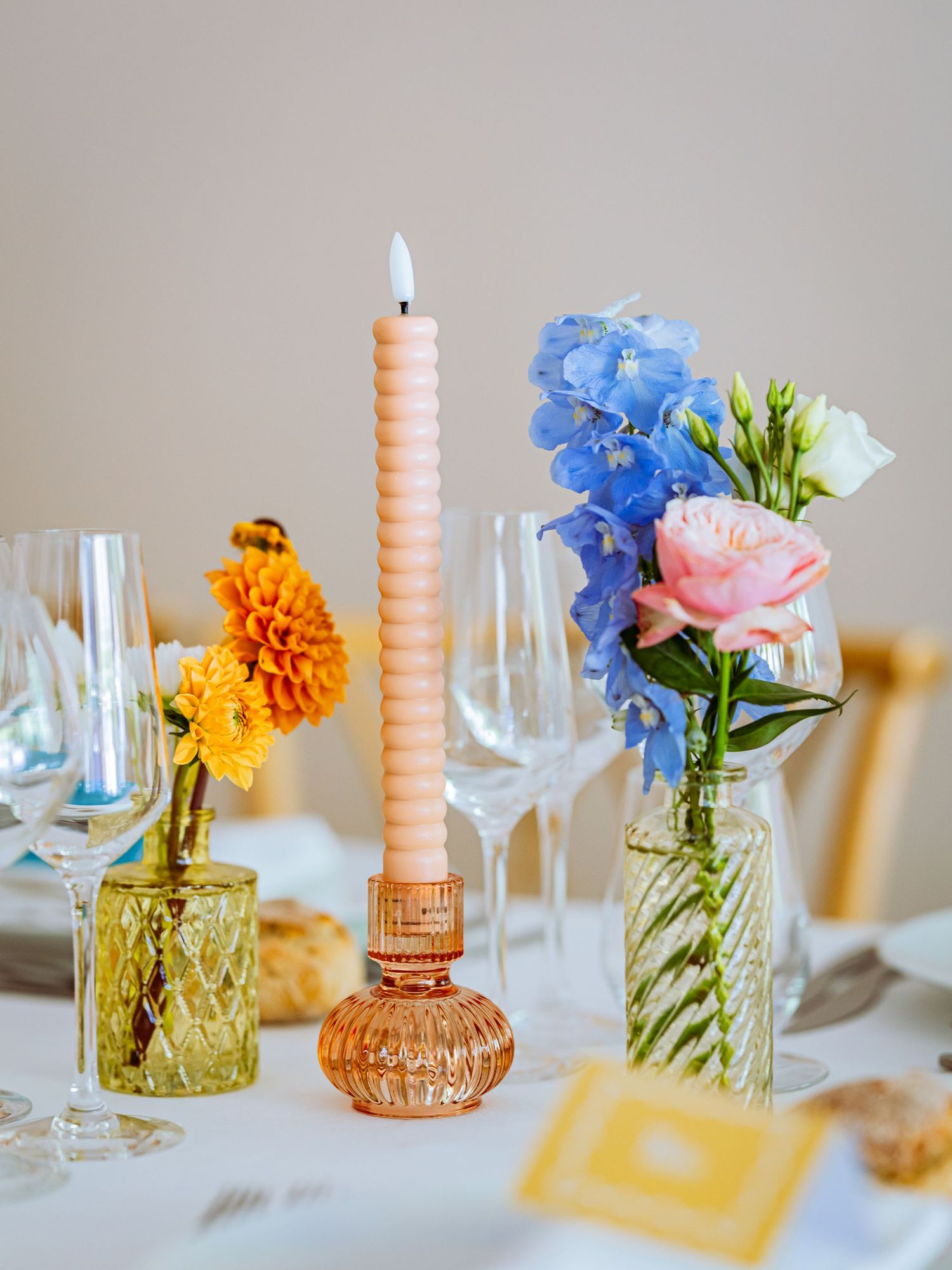 A table setting with orange and blue flowers in glass vases alongside peach candles and wine glasses.