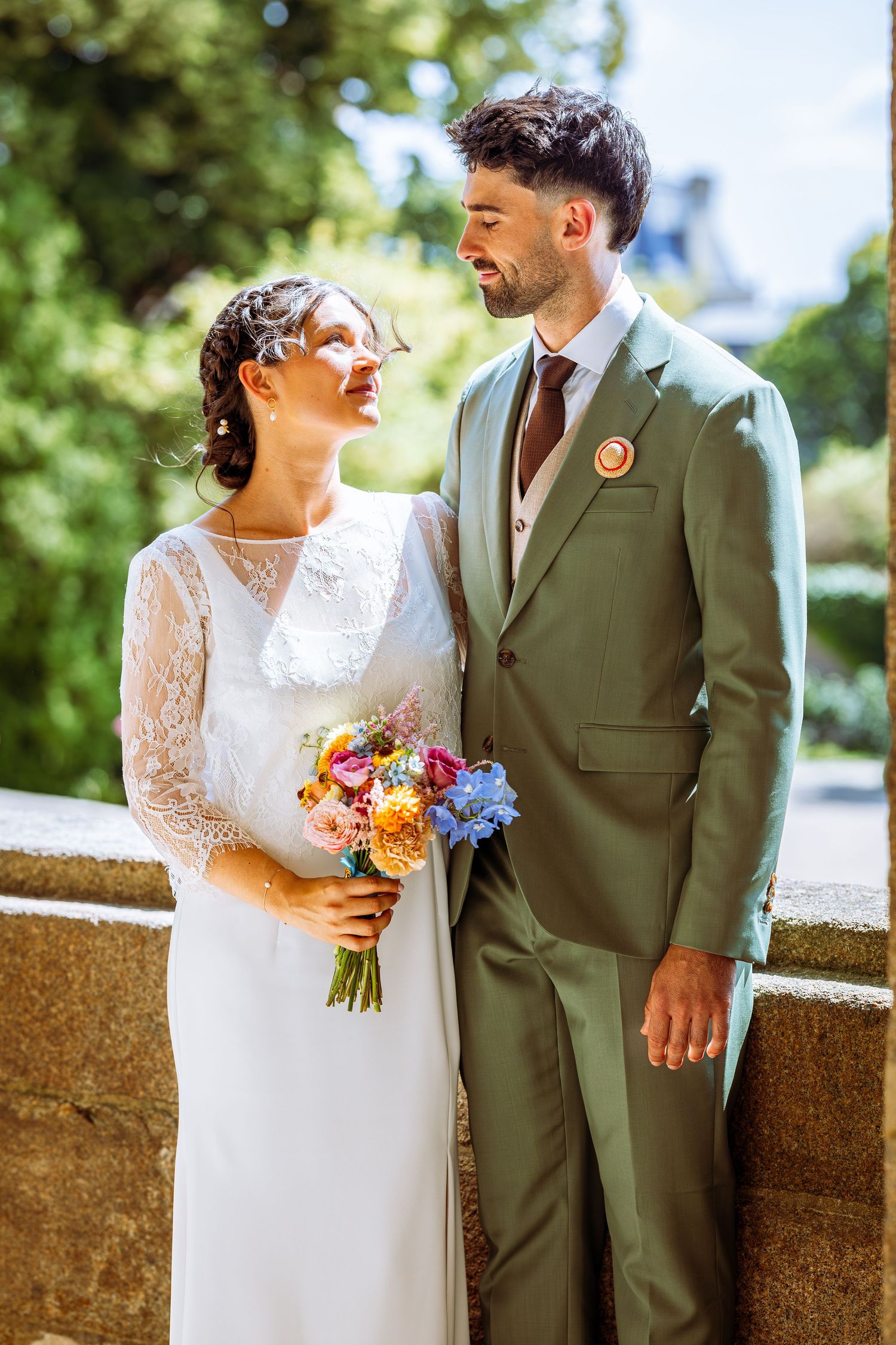 Newlywed couple in lace dress and olive suit share romantic moments against stone wall with colorful bouquet.