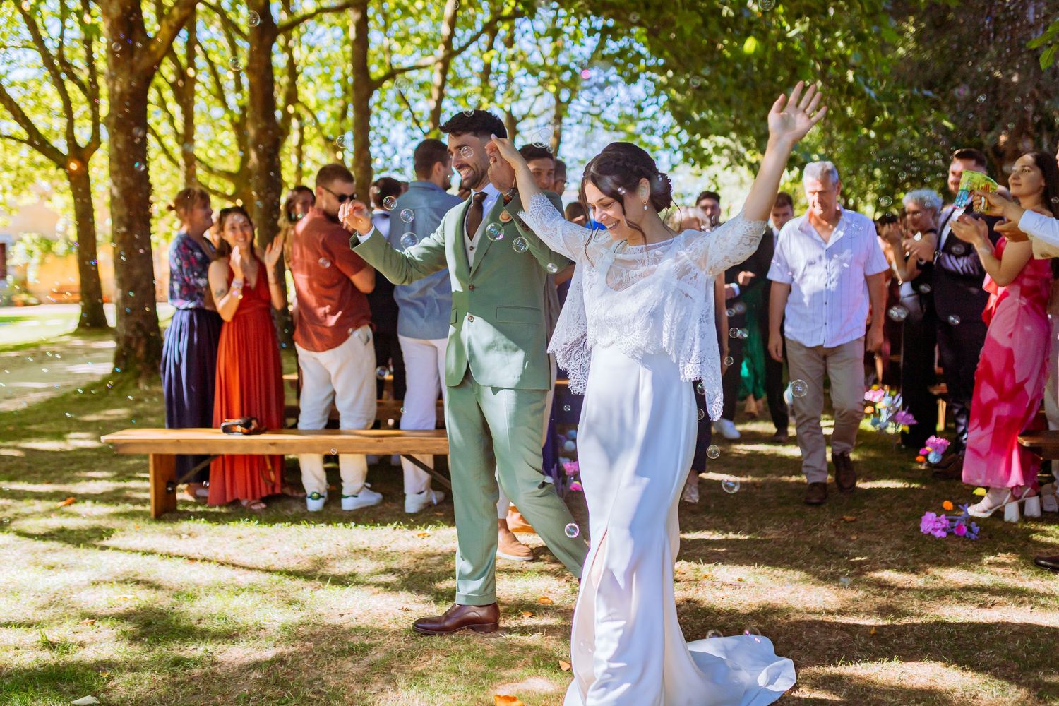 A joyful couple celebrates with raised arms during an outdoor wedding ceremony while guests cheer in the background.