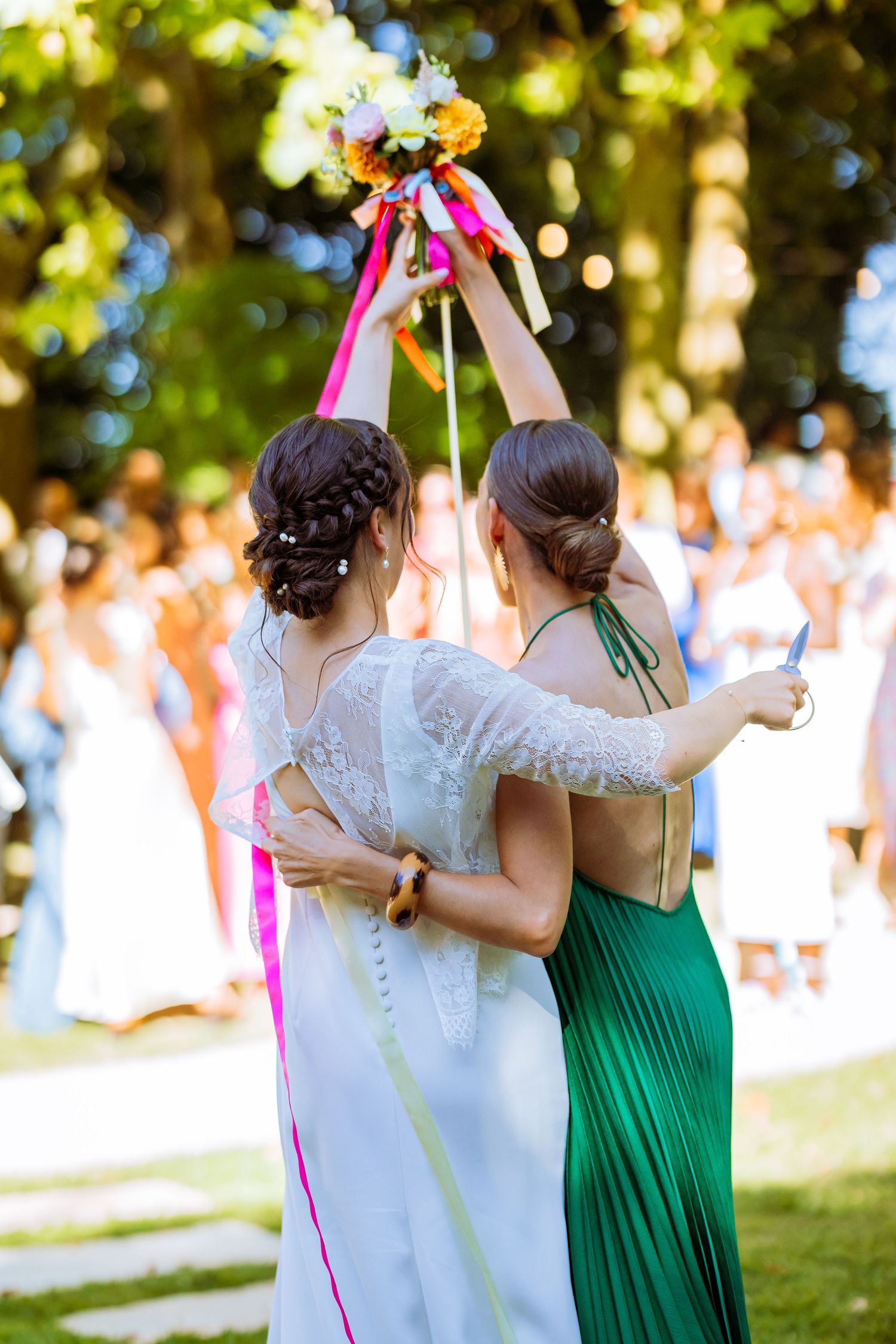 Two people in wedding and green dresses hold up a floral bouquet with colorful ribbons during an outdoor celebration.