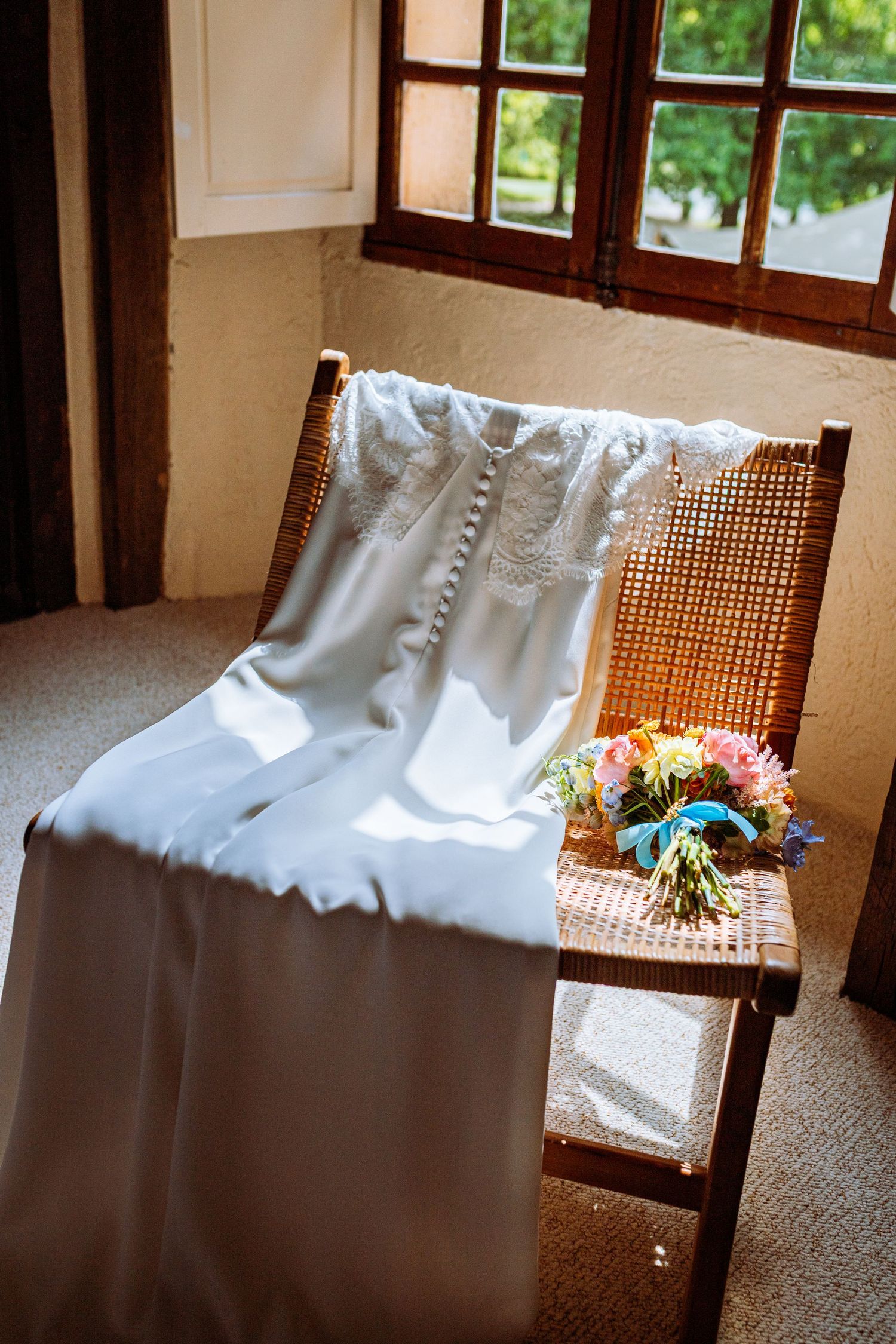 White wedding dress draped over a vintage cane-back chair with delicate pink flowers in the background.
