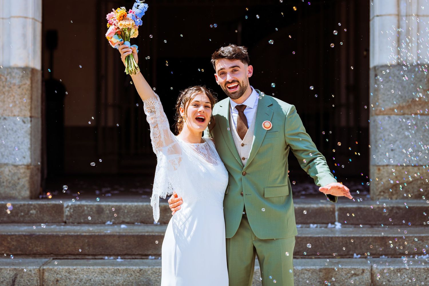 A wedding couple celebrates with confetti falling around them at a stone building venue.