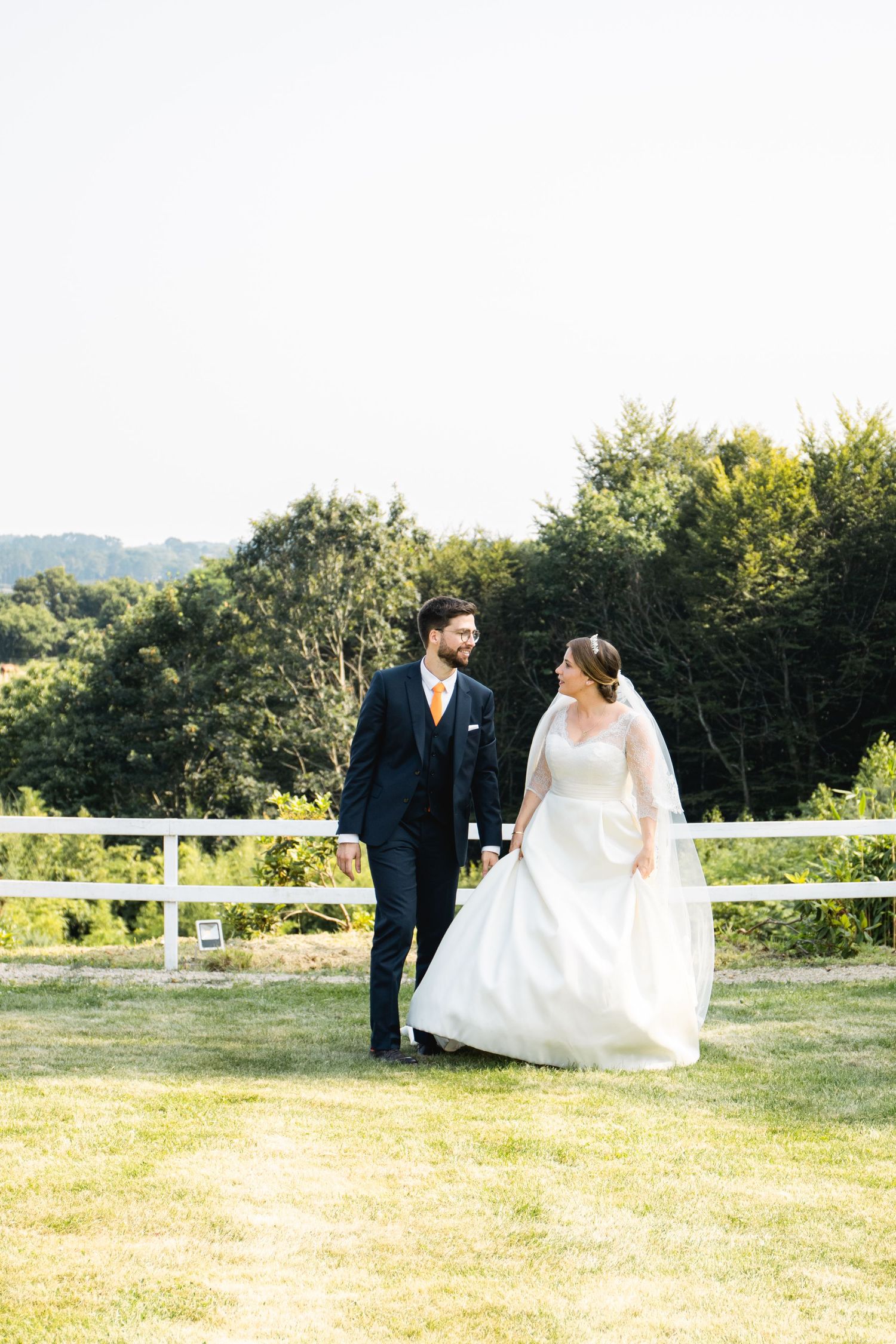 A wedding couple poses in formal attire on a sunny lawn with white fencing in the background.