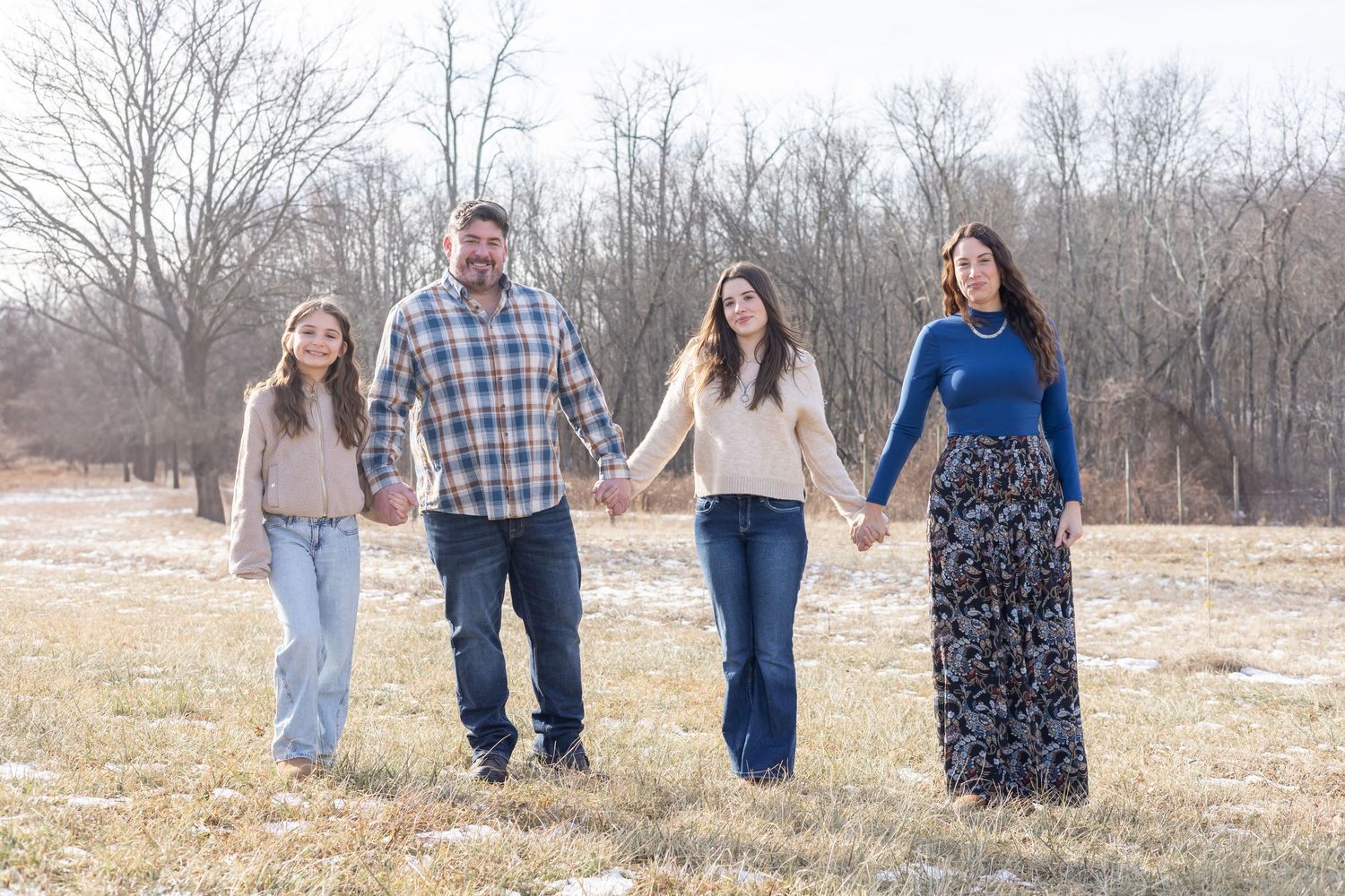 A family holds hands while standing together in a winter field with bare trees in the background.