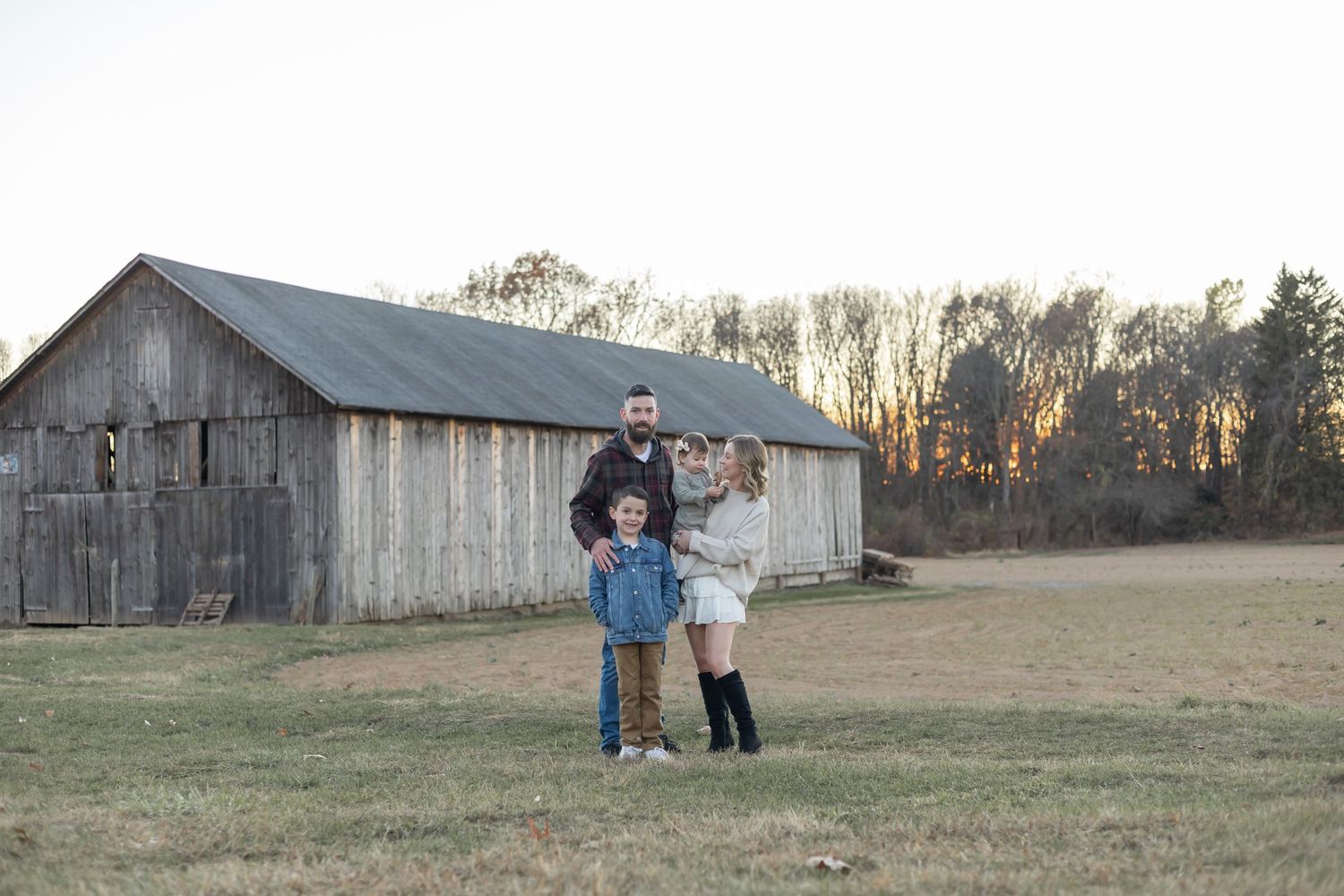 Three people stand together in front of a weathered wooden barn at sunset in a grassy field.