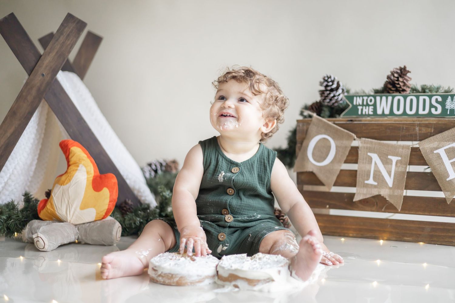 A toddler in a green romper sits on the floor next to a decorative teepee and woodsy camping-themed decor.