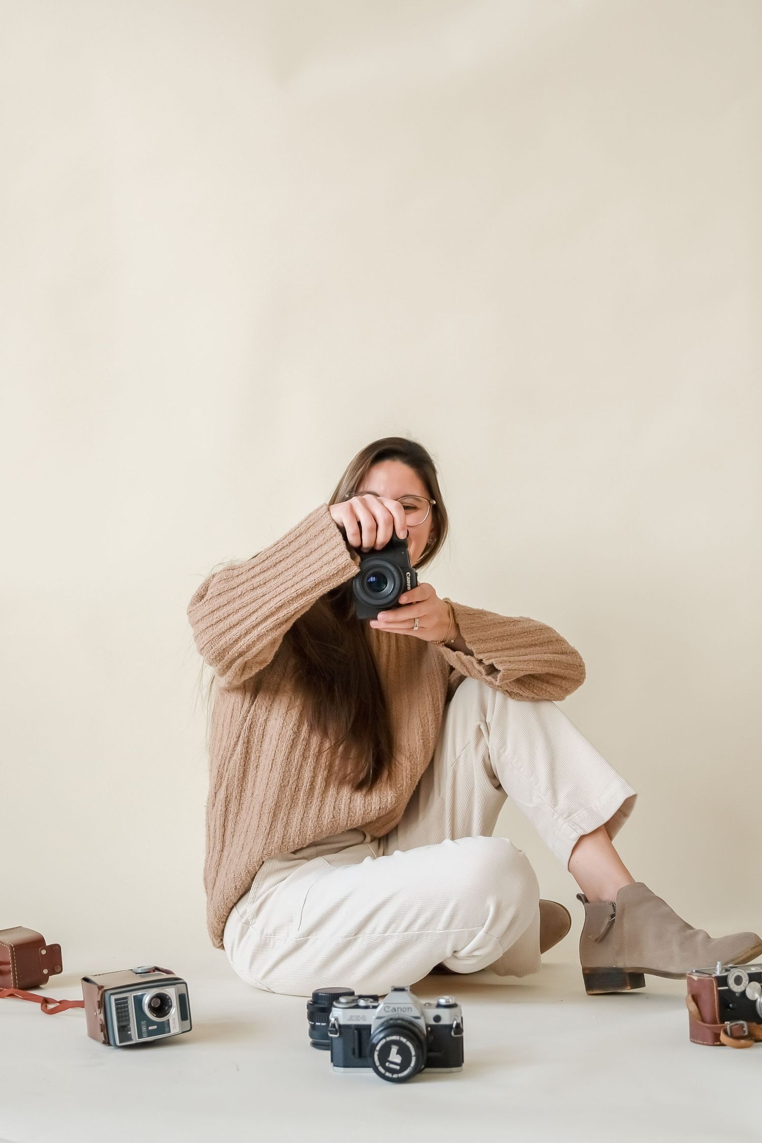 Photographer wearing beige sweater and white pants sits on floor with vintage cameras against neutral background.