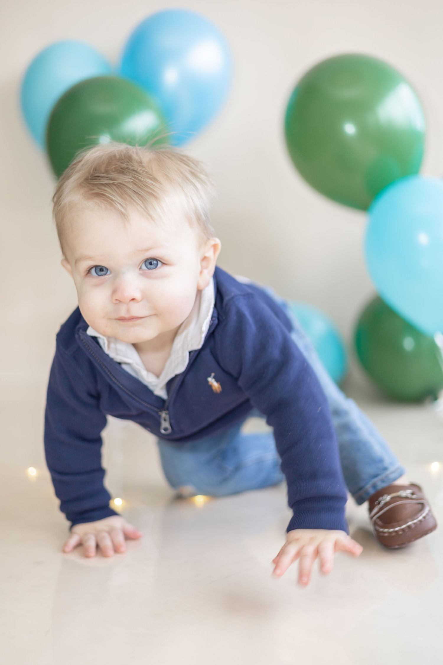 Baby crawls on floor wearing navy sweater with blue and green balloons in background during photo session.