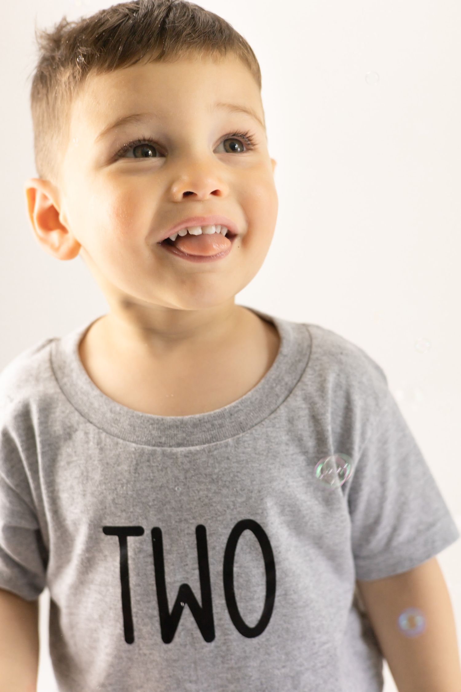 A young child wearing a gray t-shirt with the word 'TWO' written on it smiles while looking up.