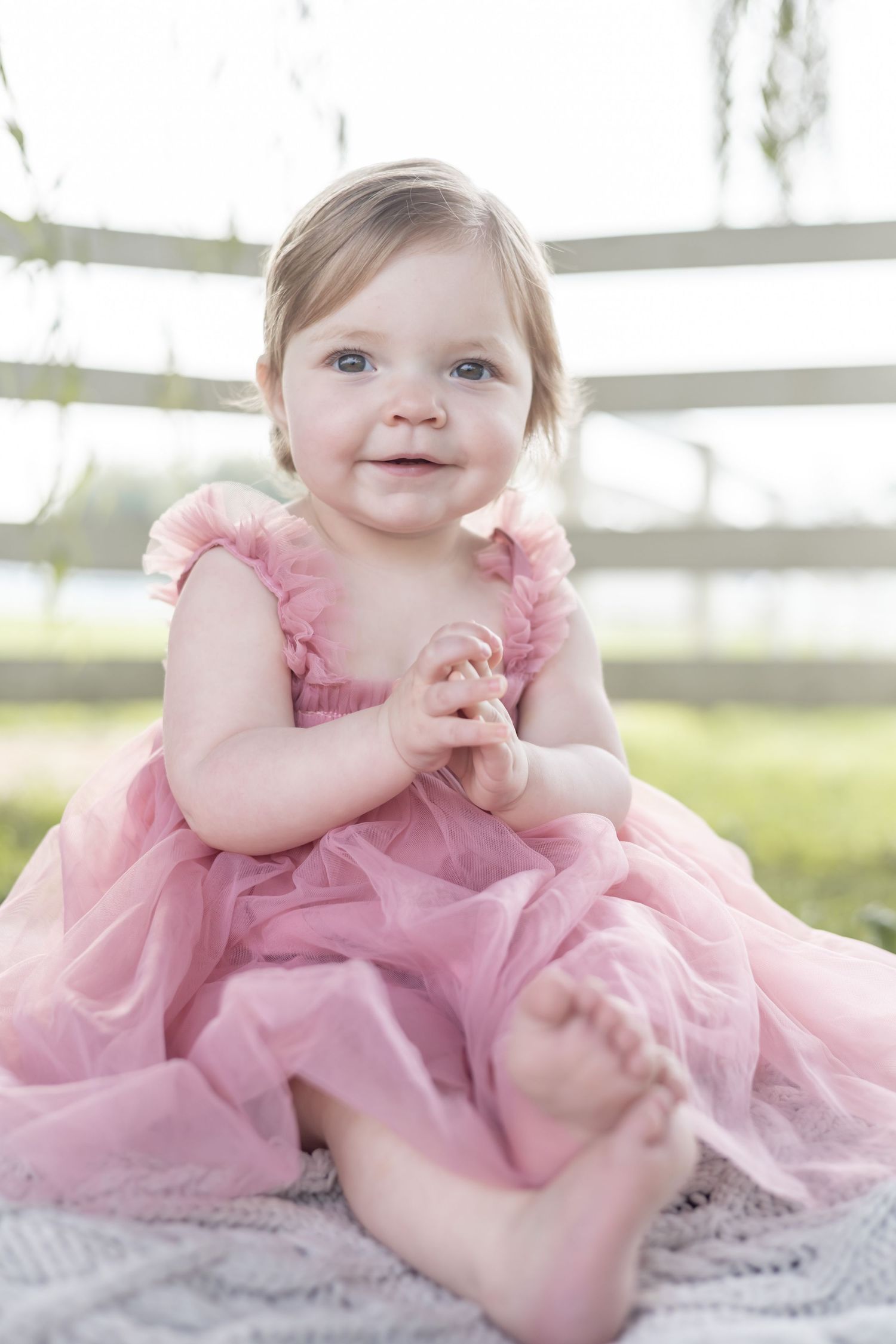 A baby in a pink tulle dress sits on a blanket outdoors, with soft sunlight filtering through trees in the background.