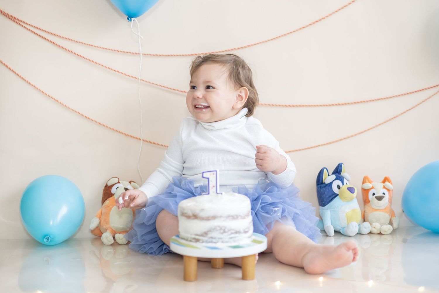 Baby sits on wooden floor with blue balloons and stuffed animals during first birthday photo shoot celebration.