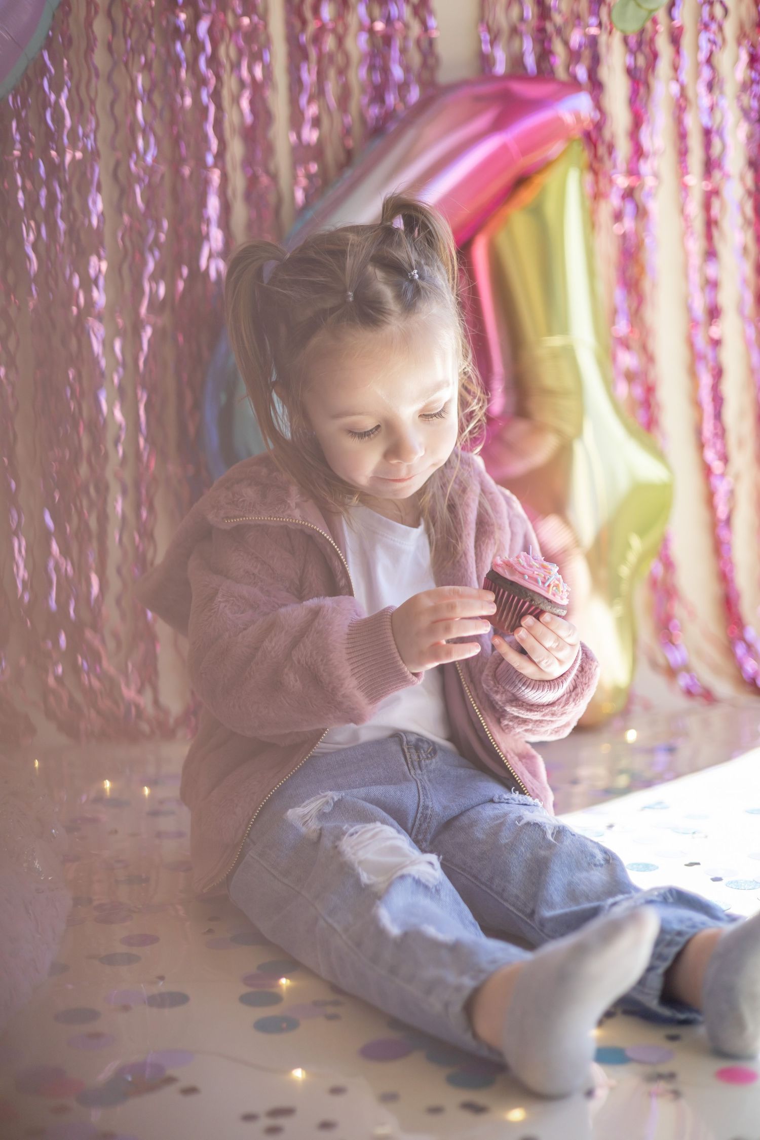 Young child in gray sweater sits on floor with sparkly backdrop and confetti, enjoying a cupcake at a party celebration.