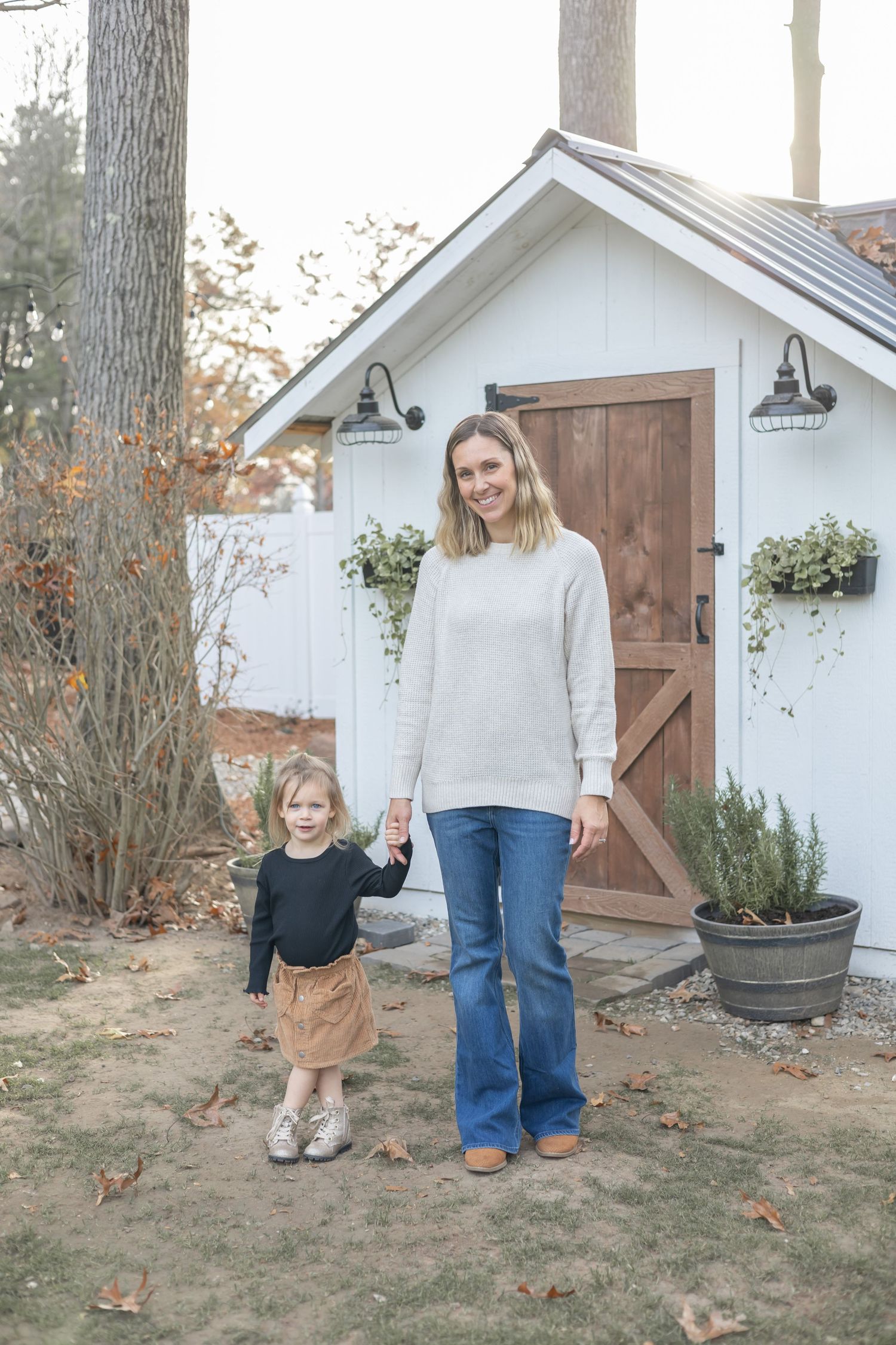 Mother and daughter walk through autumn leaves in front of a charming white garden shed with brown wooden door.