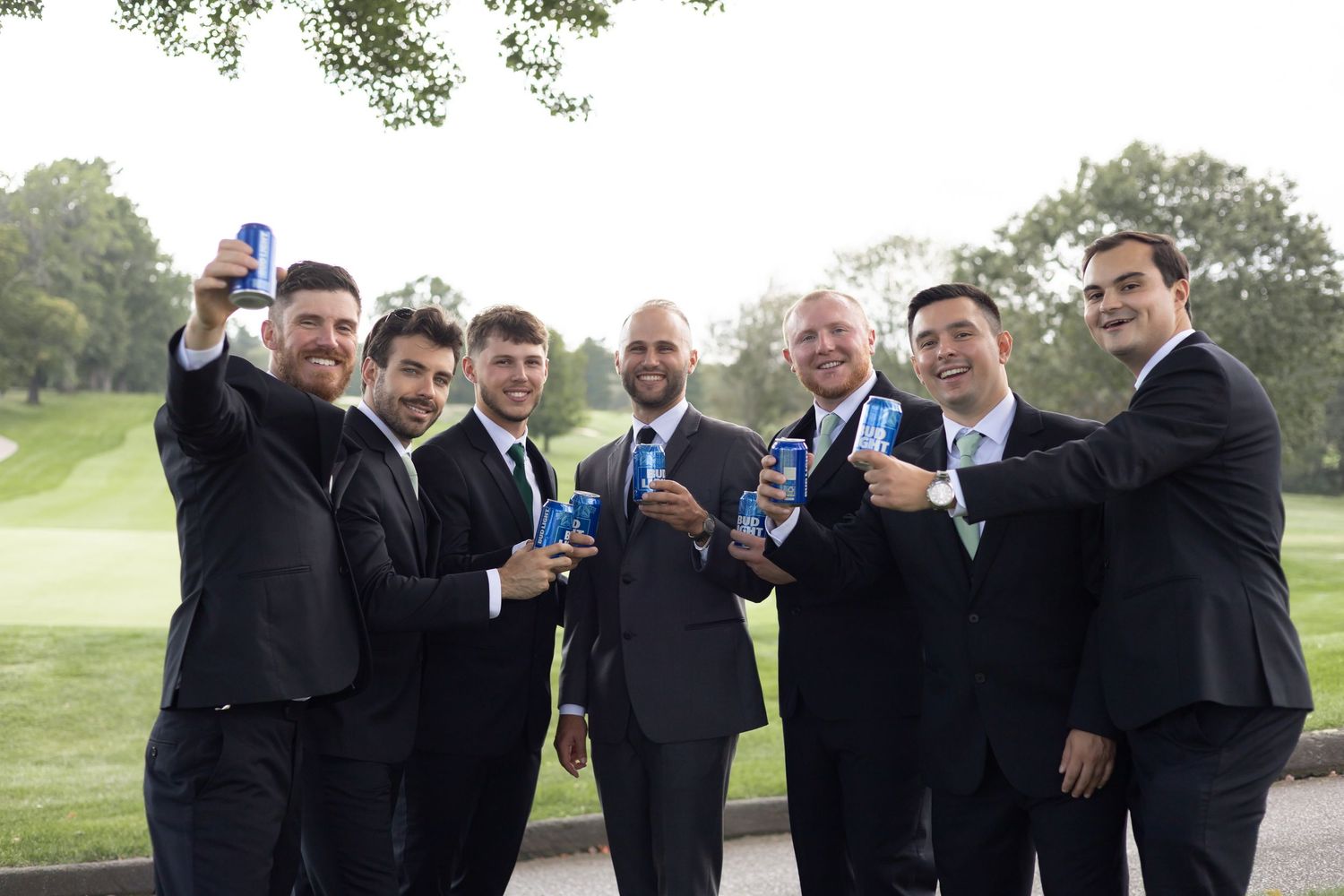 Groomsmen in dark suits raise blue drinks for a celebratory toast on a golf course.