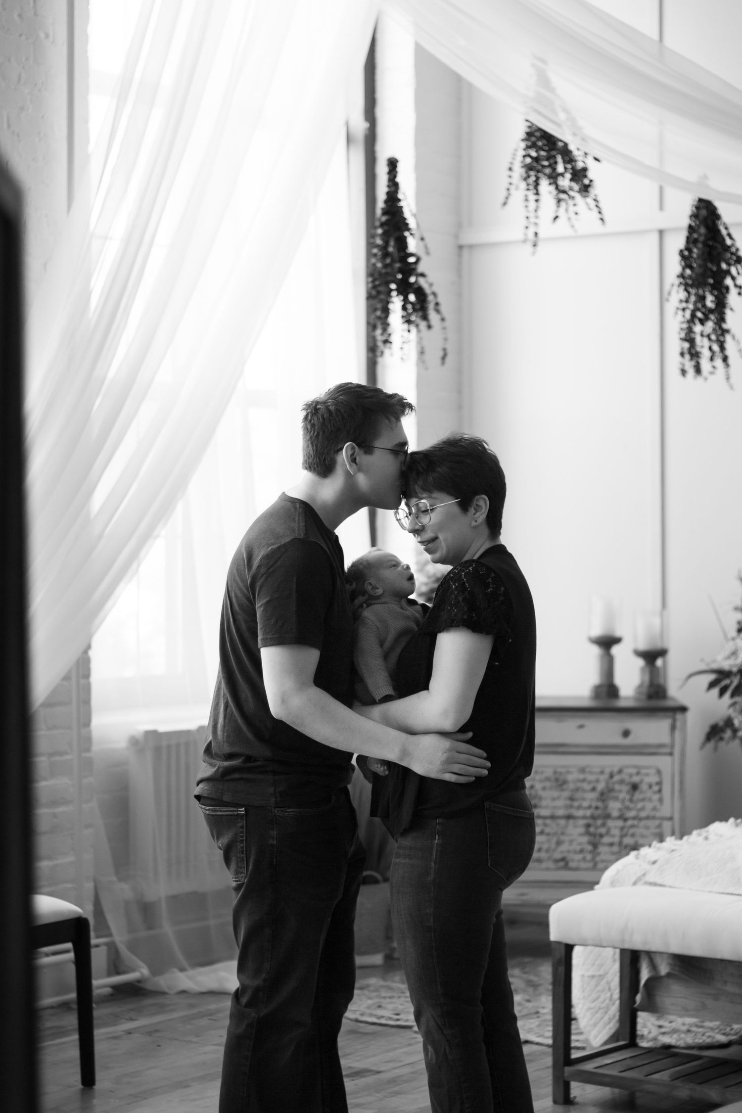 A romantic couple in black attire embrace in an intimate moment near sheer curtains and hanging plants in a bright room.