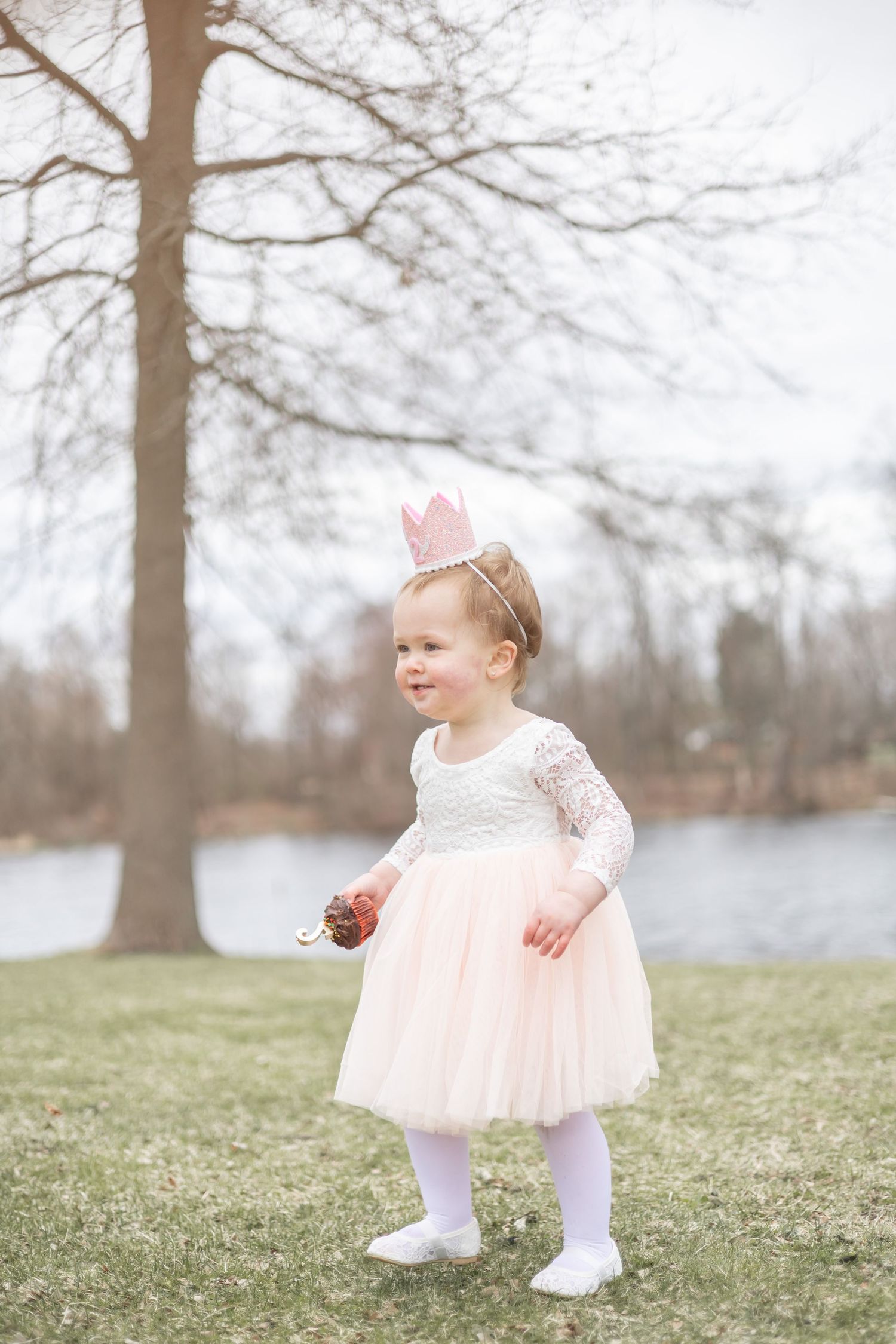 A small child in a white lace and pink tulle dress with a crown stands on grass near a lake in winter.