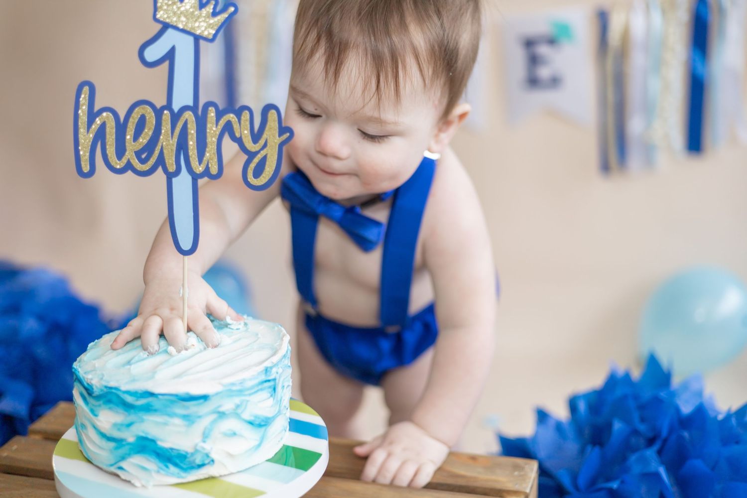 Toddler in blue suspenders reaches for a white and blue birthday cake with decorative marbling pattern.
