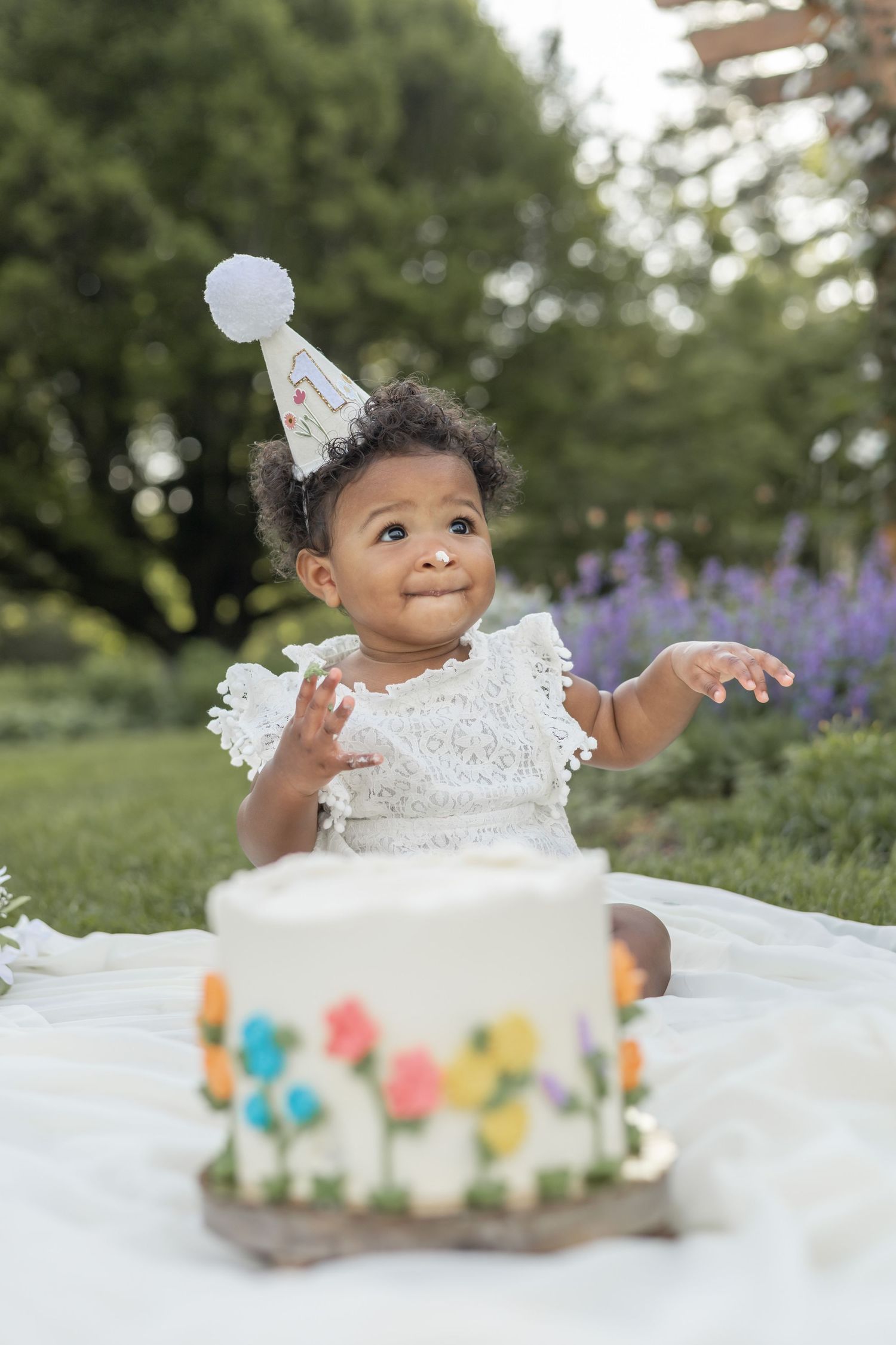 Sweet first birthday celebration outdoors with white cake decorated with colorful flowers and tiny party hat.