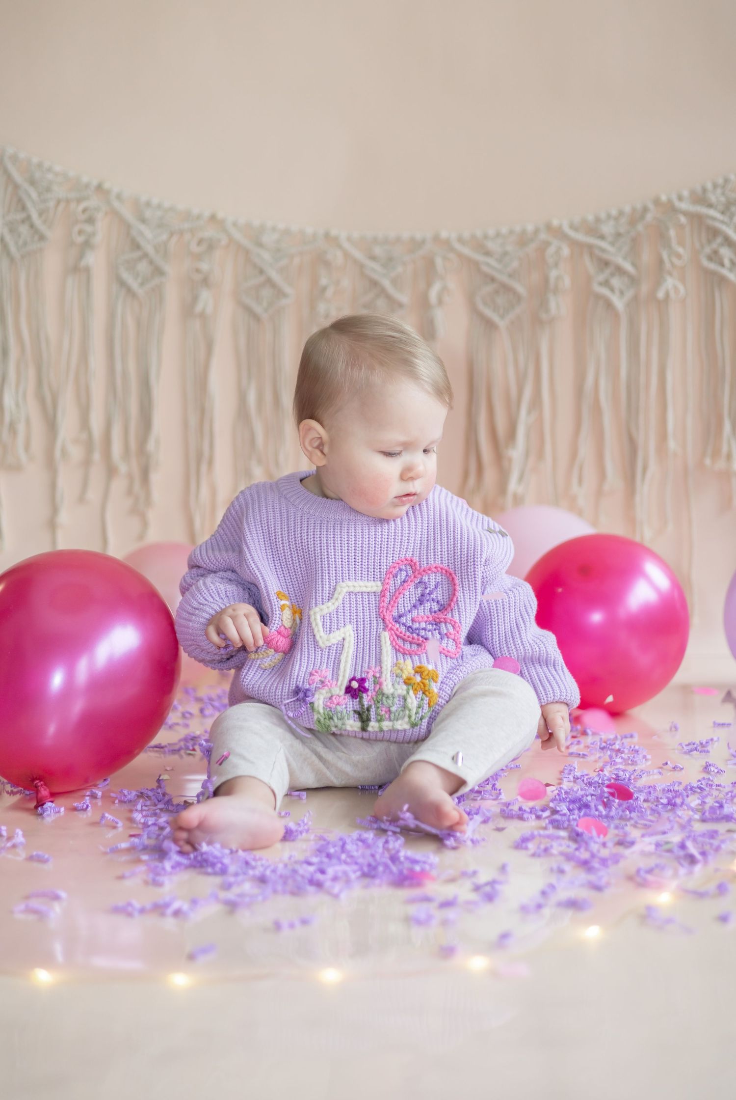 Baby in purple sweater sitting with pink balloons and confetti on beige backdrop with twinkle lights and streamers.