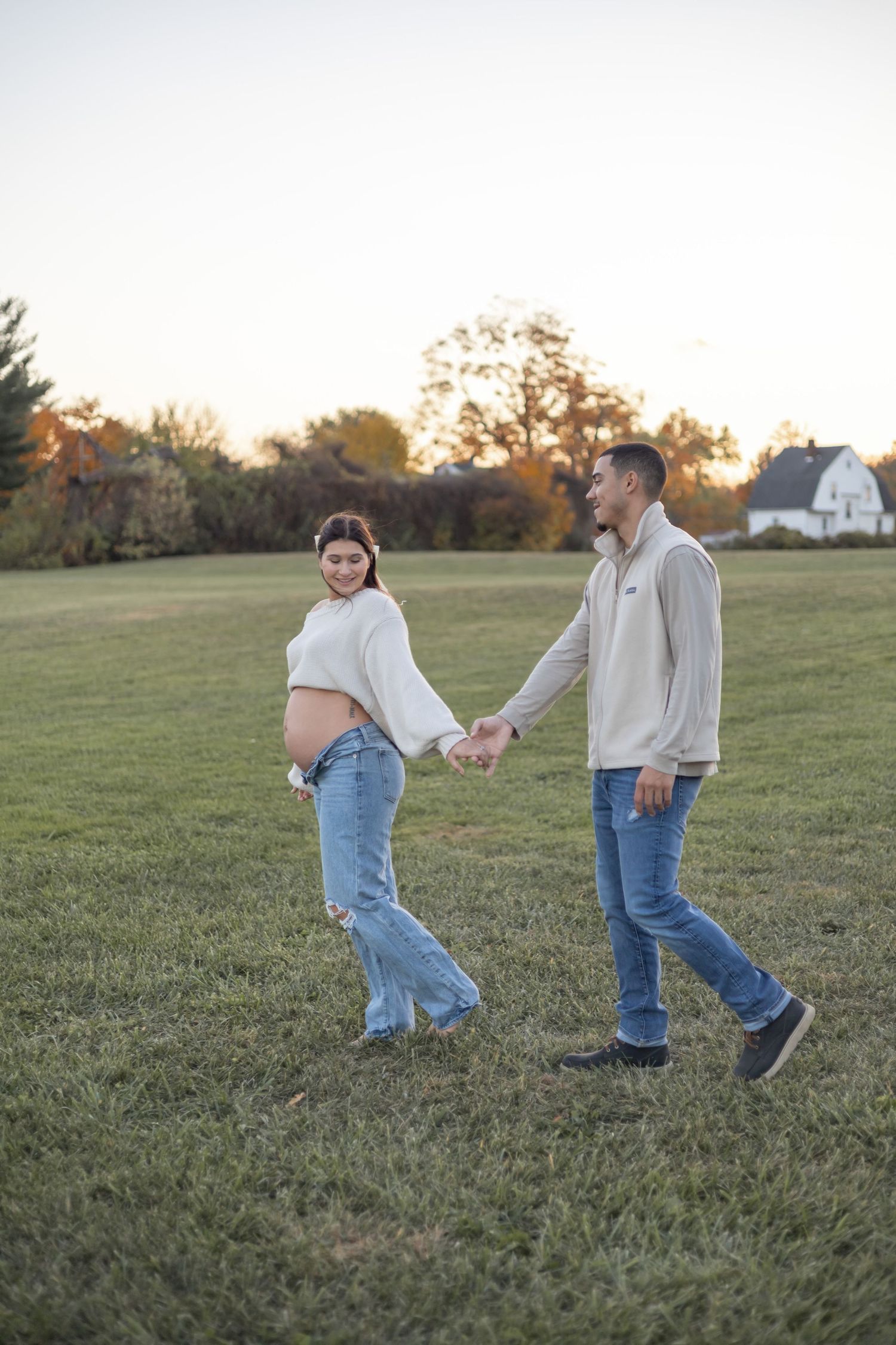 Couple holds hands and walks through a grassy field at sunset wearing jeans and light-colored tops.
