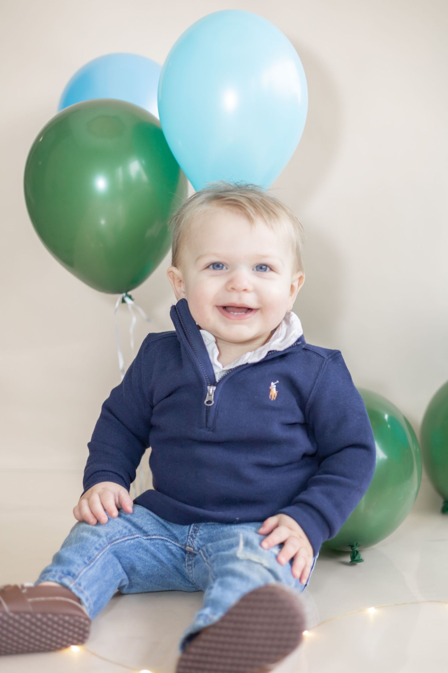 A baby in a navy blue sweater sits on the floor with blue and green balloons in the background.
