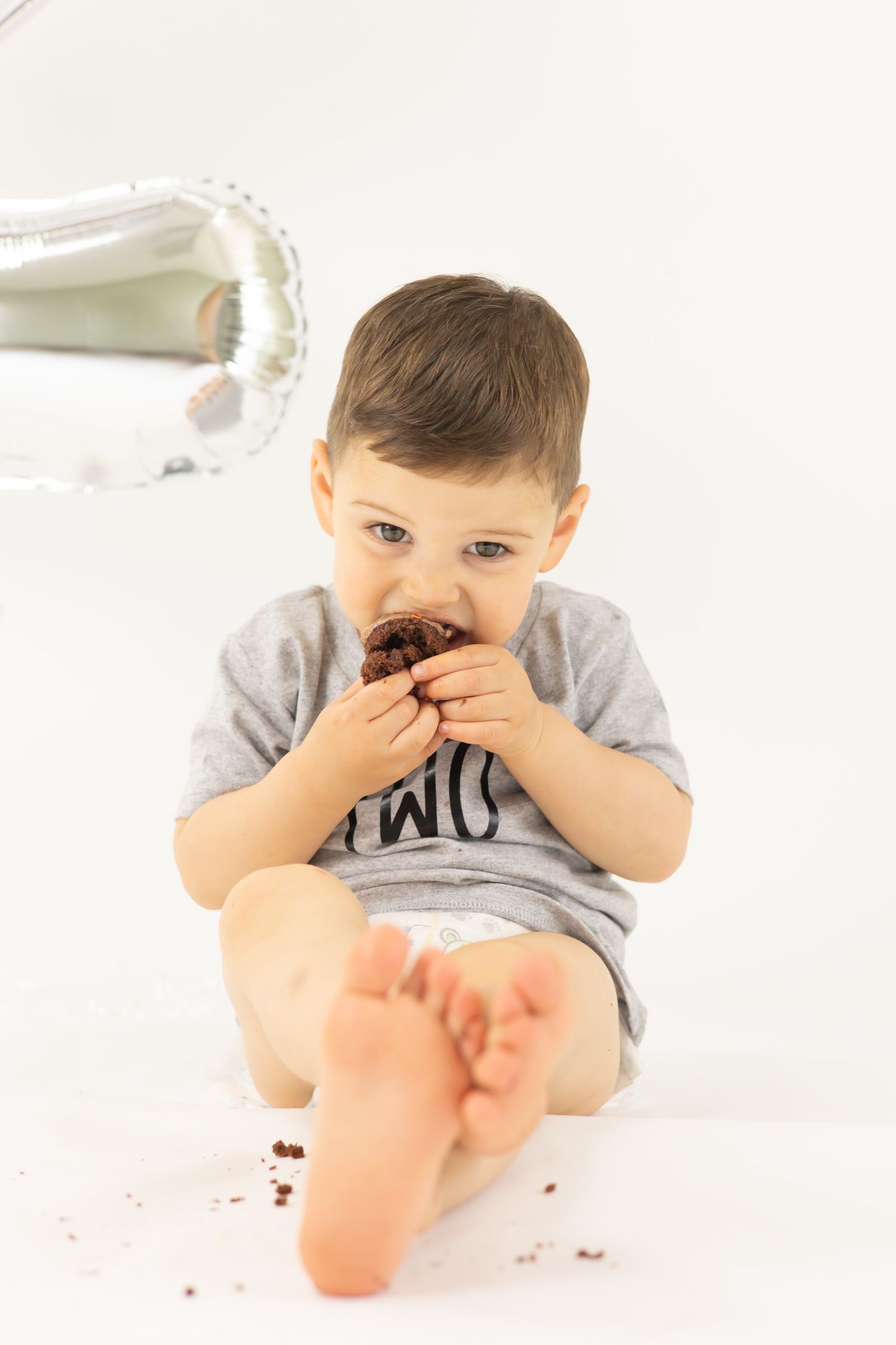 A child in a gray t-shirt lays on a white surface and giggles while covering their mouth with their hands.